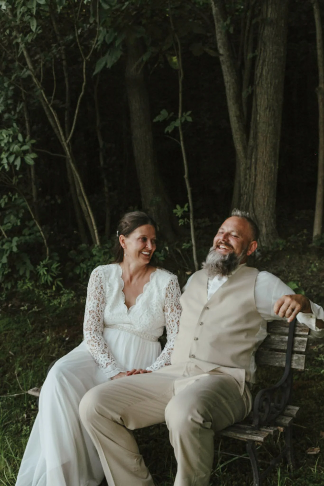 A woman in a white lace dress and a man in a beige vest and trousers sitting on a park bench, smiling and laughing, surrounded by trees and greenery.