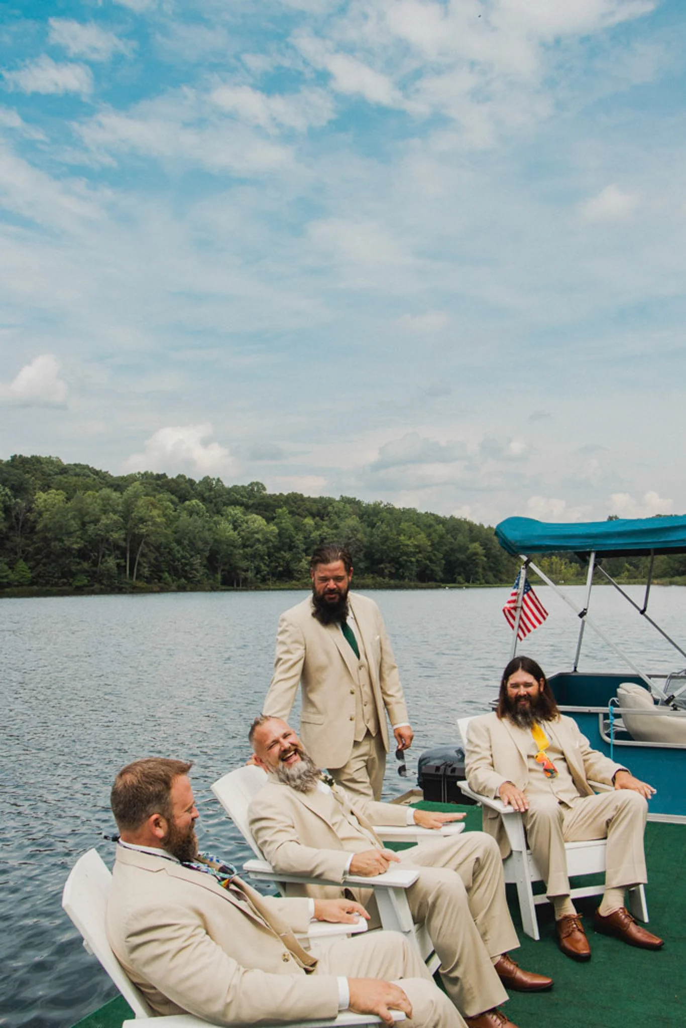 Four men in beige suits relaxing and smiling on a boat by a lake, with a background of trees and a partly cloudy sky.