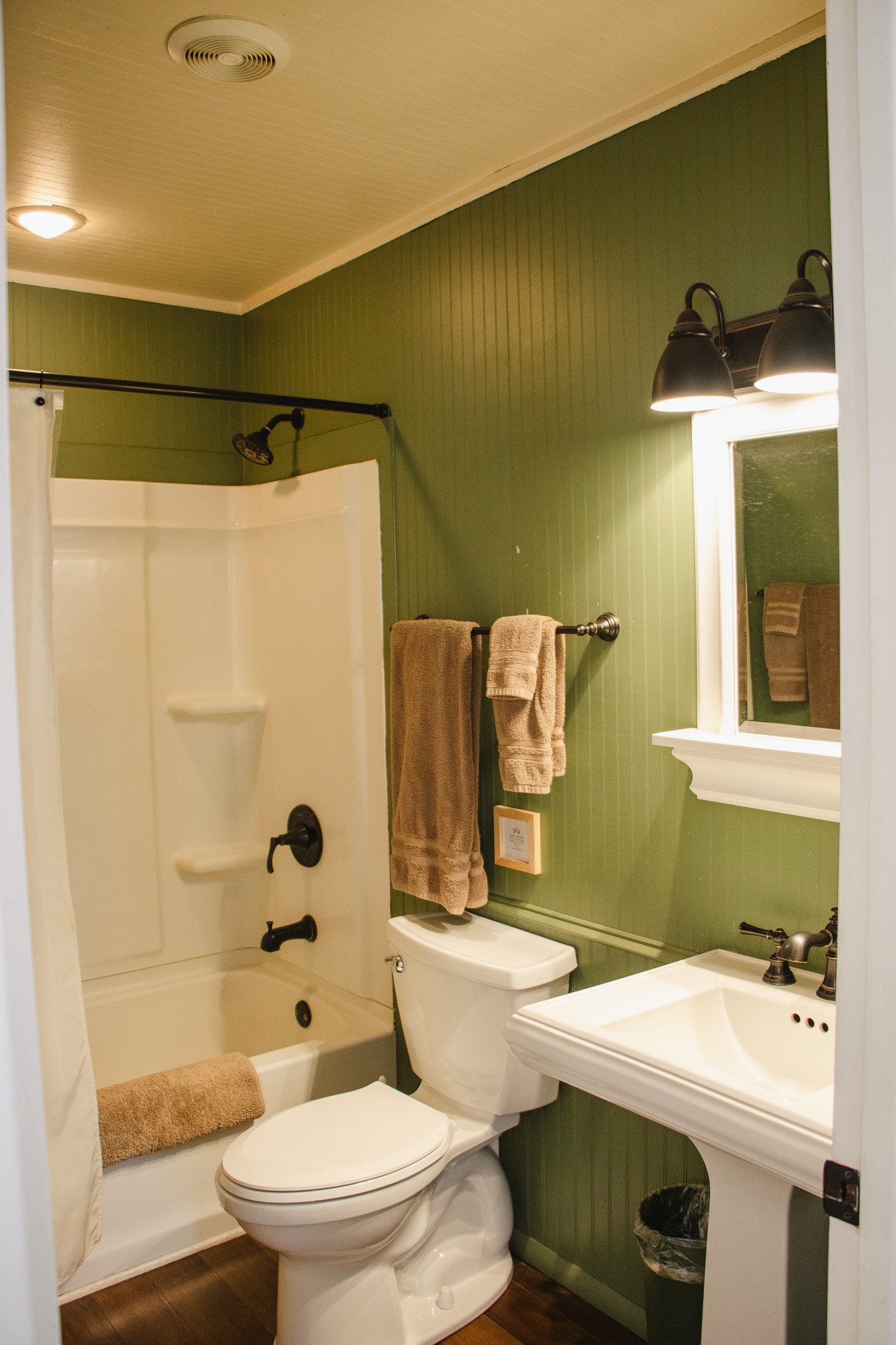 A small bathroom with green paneled walls, a bathtub with a shower, a white toilet, a white sink, a mirror, and beige towels hanging on a towel rack.