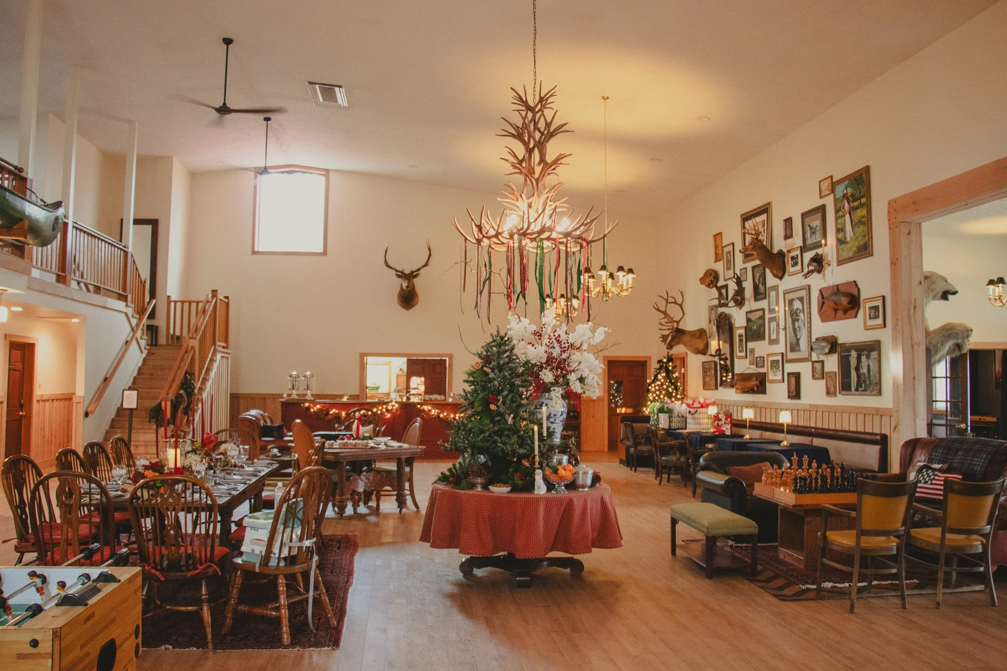 A large, decorated room with wooden floors and walls, featuring a central Christmas tree in front of a round table. The room has wall-mounted deer and animal head displays, and various framed pictures. A rustic chandelier made of antlers hangs from t