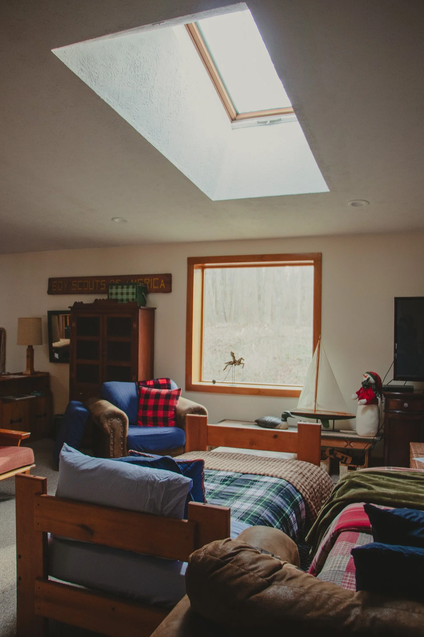 A cozy living room with a skylight and a large window, decorated with a boat model and stuffed snowman, and furnished with sofas, armchair, and wooden furniture, with a 