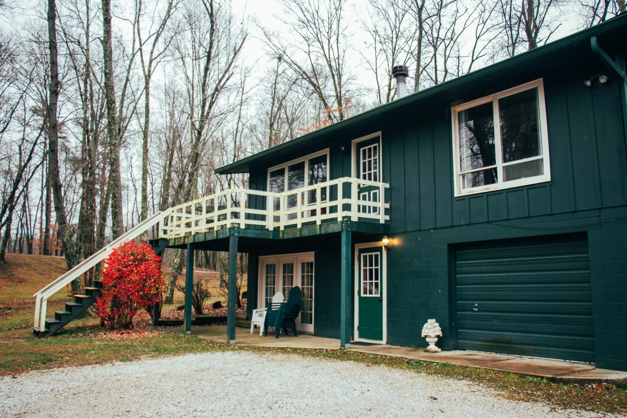A two-story dark green house with a white deck and stairs, surrounded by leafless trees and a gravel driveway.