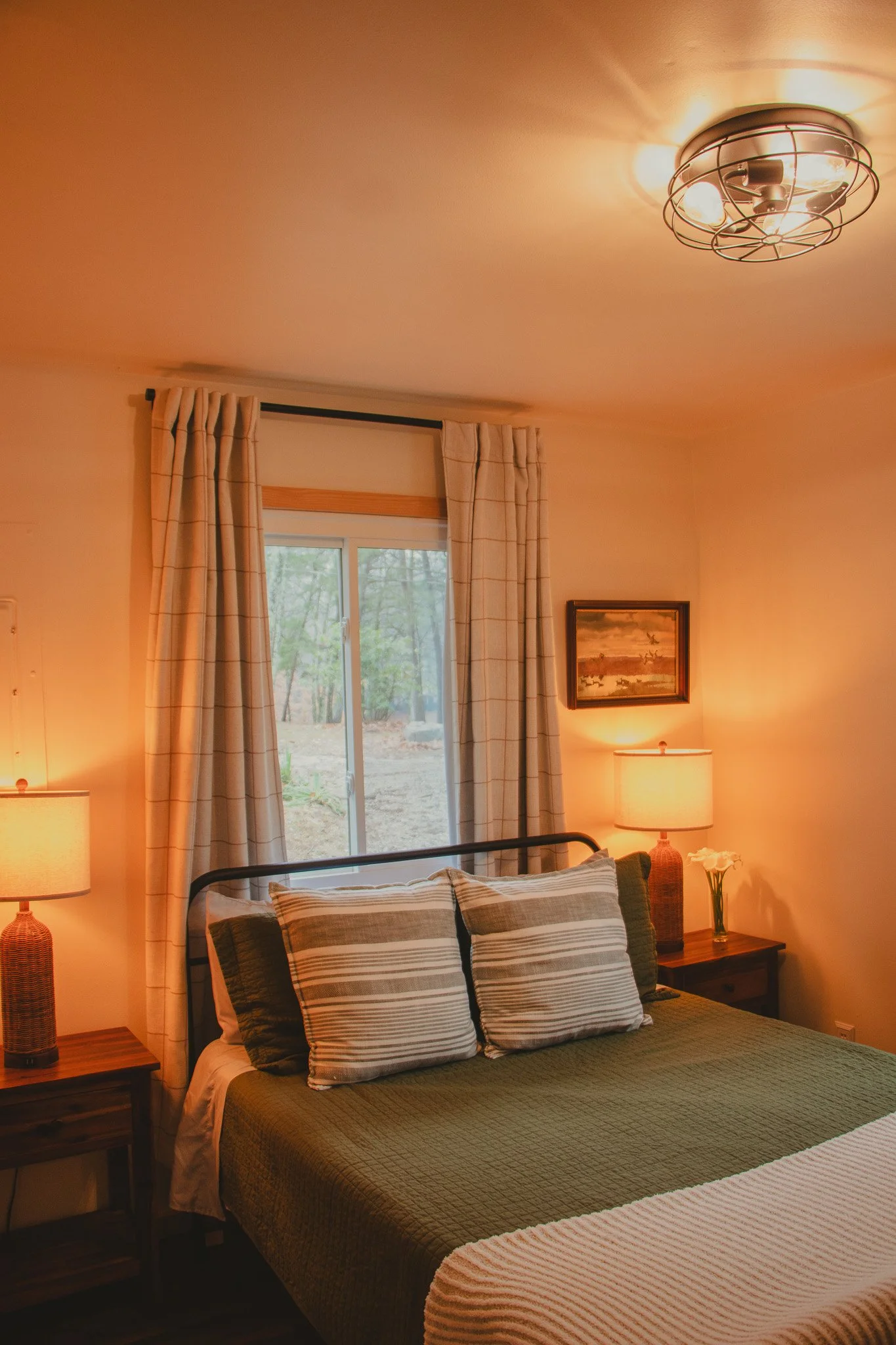 A cozy bedroom with a large bed, two bedside lamps, a window with curtains, framed artwork, and a floral arrangement.