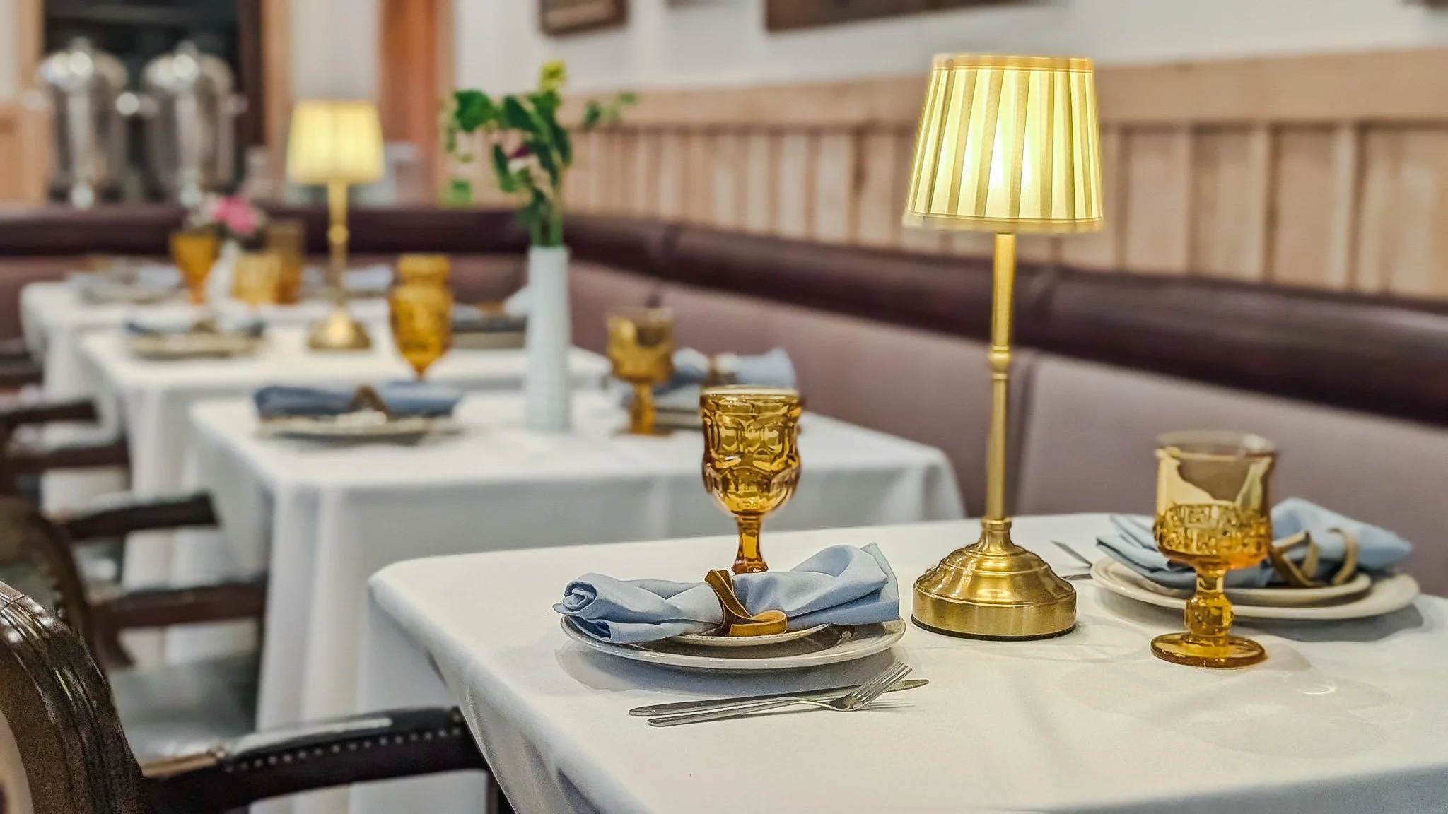 Set dining table with white tablecloth, gold-rimmed glasses, blue napkins, silverware, a gold table lamp, and floral centerpieces in a restaurant.