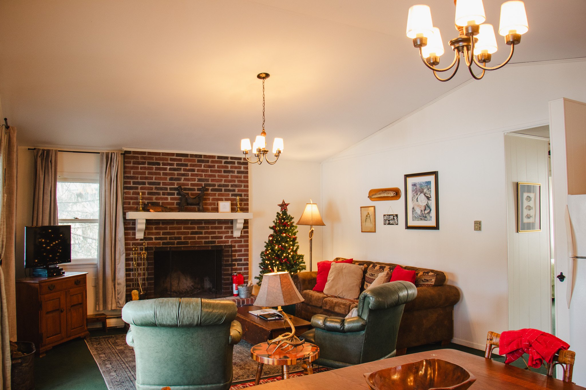 Living room decorated for Christmas with a Christmas tree, sofa with red and beige pillows, green armchair, brick fireplace, and chandelier lighting.