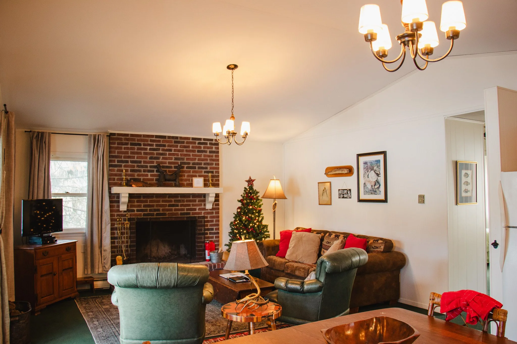 Living room decorated for Christmas with a Christmas tree, sofa with red and beige pillows, green armchair, brick fireplace, and chandelier lighting.