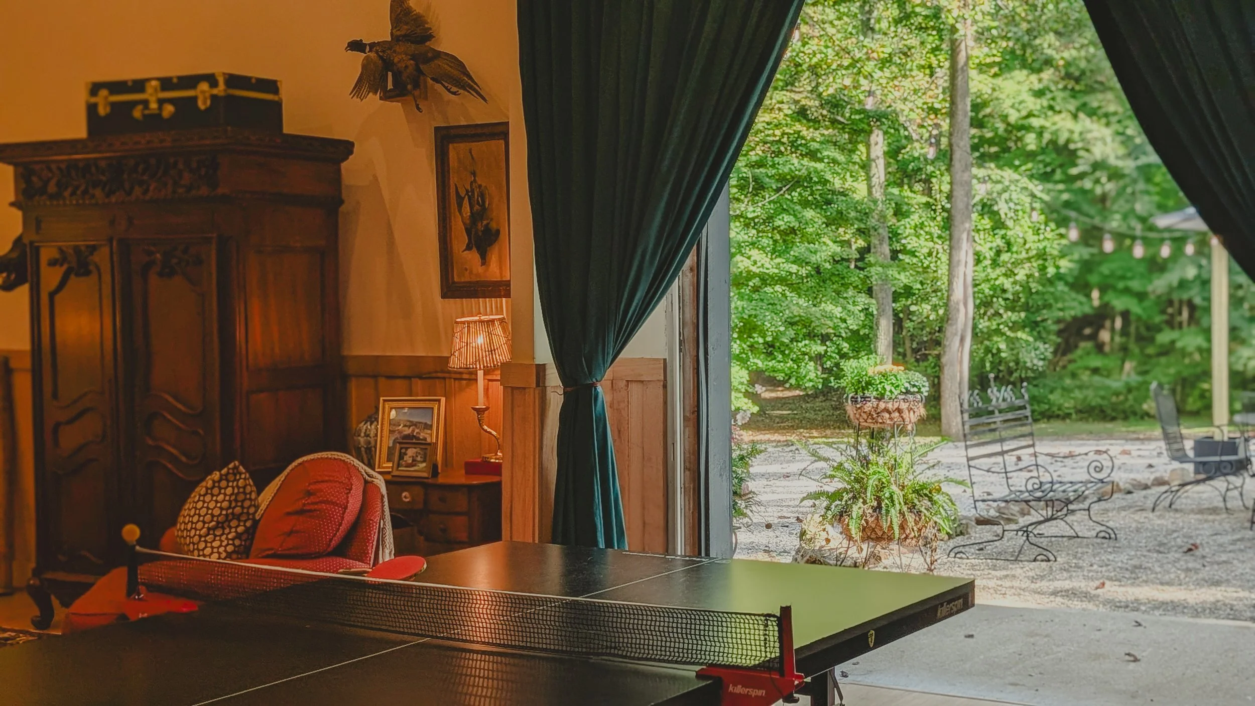 Interior of a cozy room with a ping pong table and a large open sliding door leading outdoors to a patio with trees.