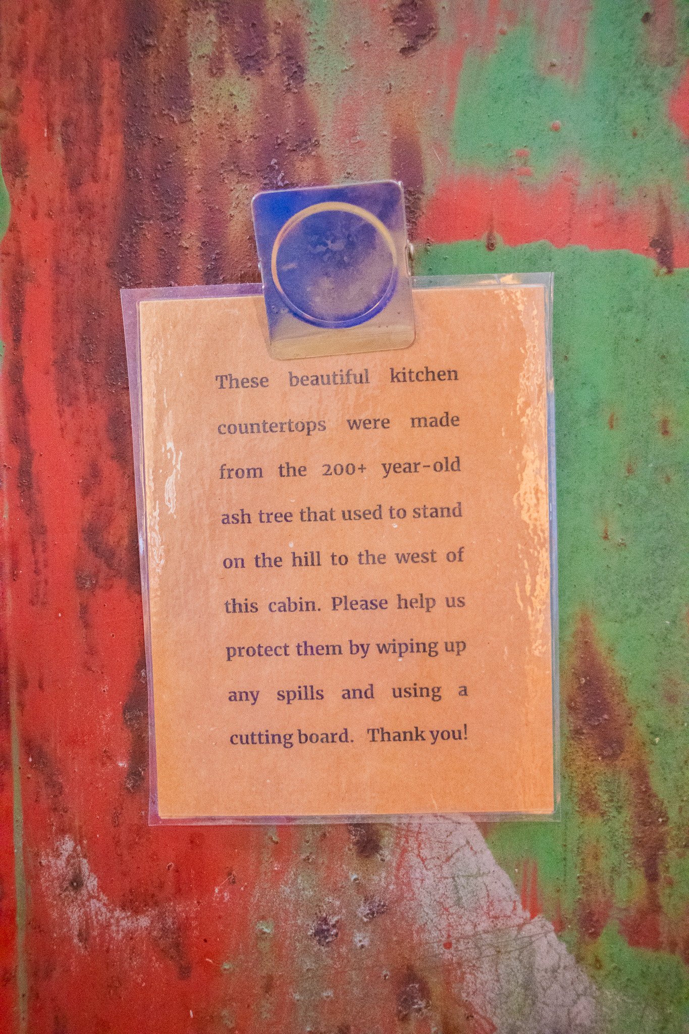 Sign on a colorful, rustic kitchen countertop explaining that the countertops are made from 200+ year-old ash tree used to stand on a hill near the cabin, asking for help in protecting them by wiping spills and using a cutting board.