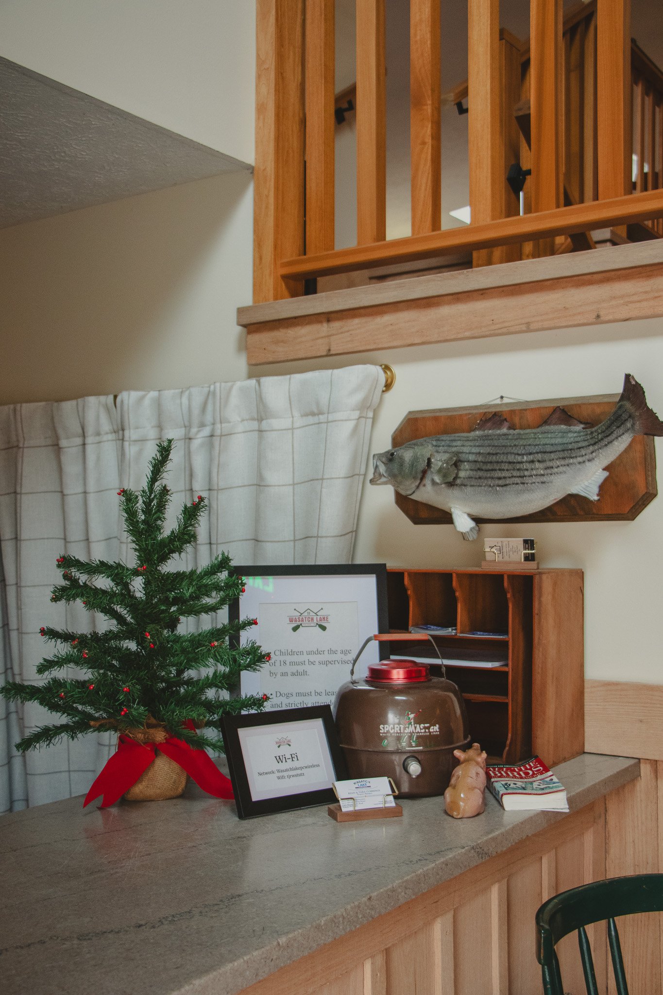 A small decorated Christmas tree with a red ribbon ribbon on a counter next to Wi-Fi and information signs, a brown thermal coffee pot, a ceramic pig figurine, and a stack of magazines. A mounted fish is on the wall, and a wooden railing and staircas