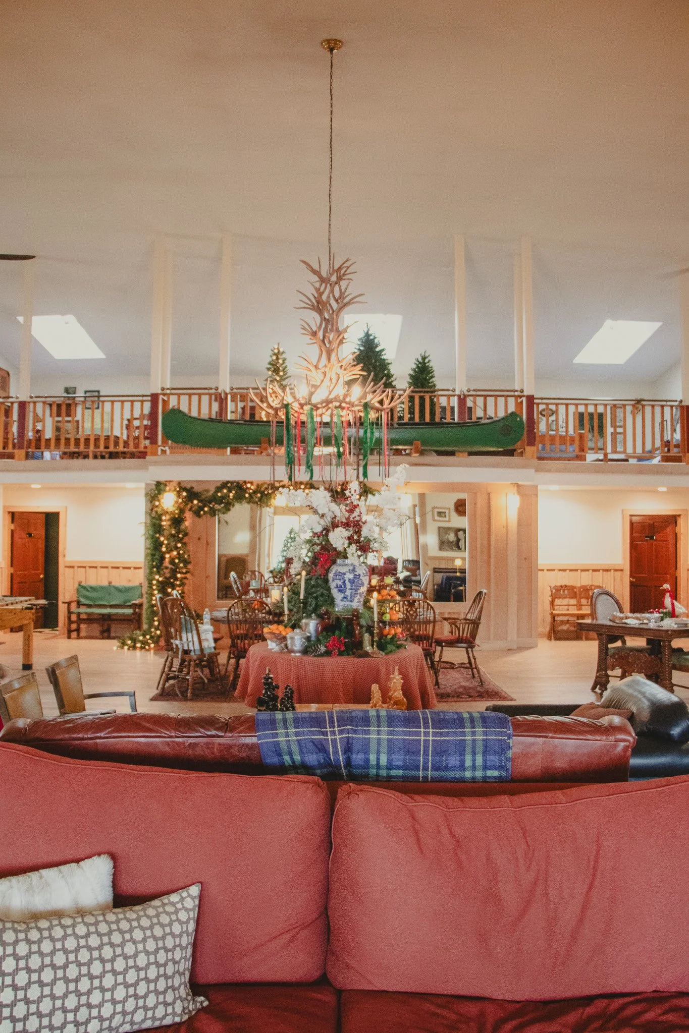 Living room decorated for Christmas with a large floral centerpiece, string lights, and Christmas decorations. A round table with a red tablecloth and various dishes. A chandelier resembling antlers hanging from the ceiling. Wooden railings and green