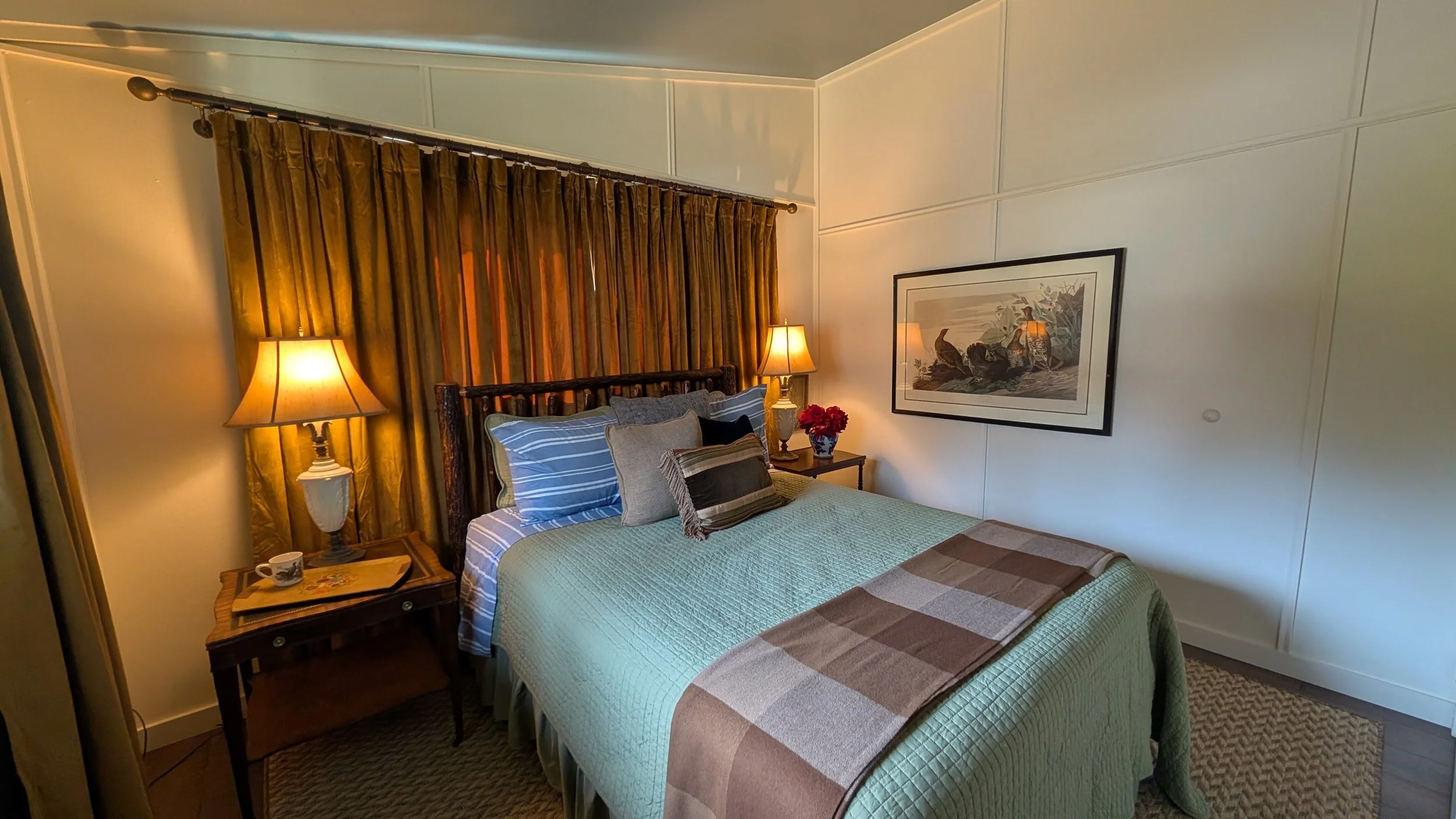 A cozy bedroom with a wooden bed frame, two pillows, a bedside table, a lamp, a framed picture, cream walls, and a window with velvet curtains.