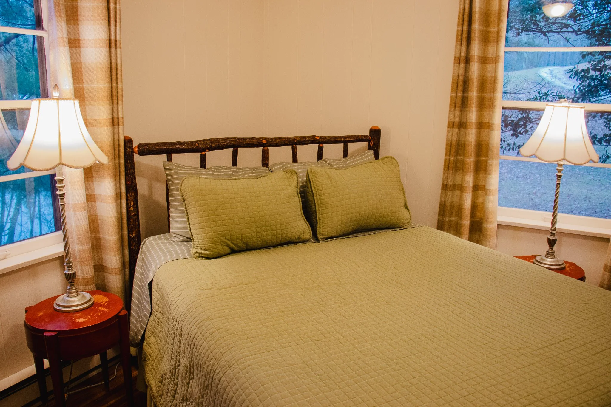 A cozy bedroom with a rustic wooden headboard, a bed with green quilted bedding and matching pillows, two side tables with lamps, and windows with plaid curtains showing trees and a winter scene outside.