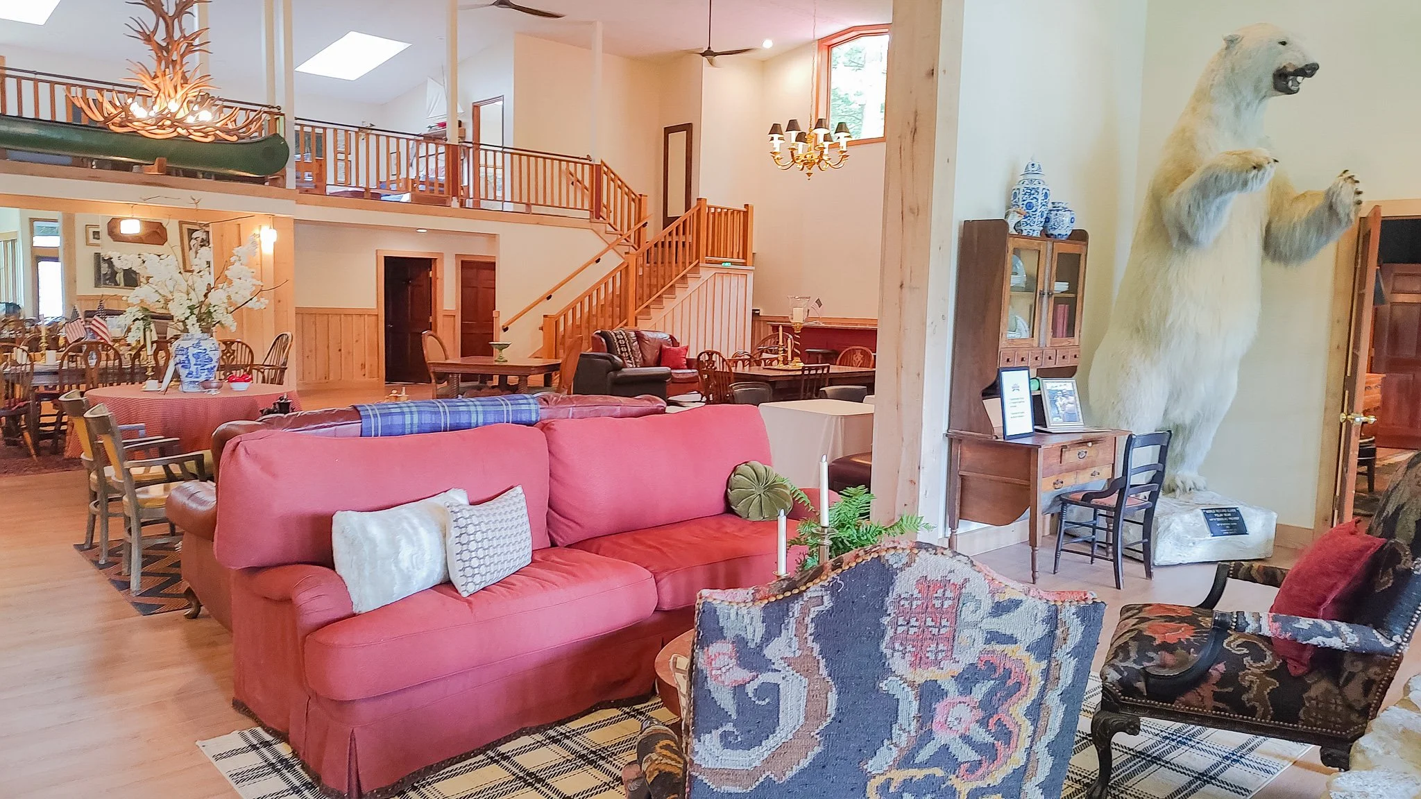 Interior of a cozy living room with wooden accents, a red sofa, patterned chairs, a dining area, and a large bear sculpture standing on its hind legs on the right side.