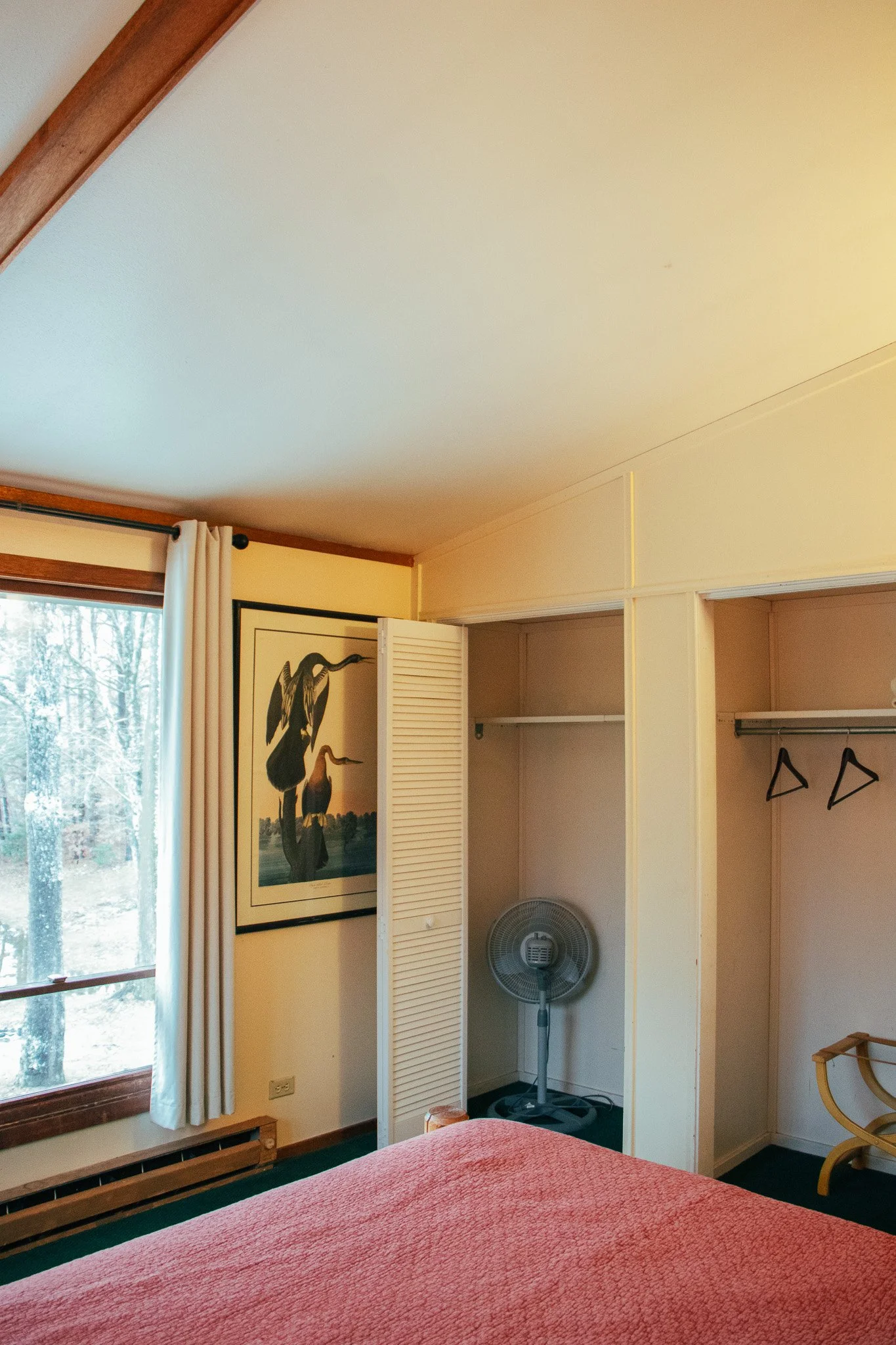 A bedroom with an open closet, a standing fan, a framed picture of herons on the wall, and a window with white curtains showing trees outside.