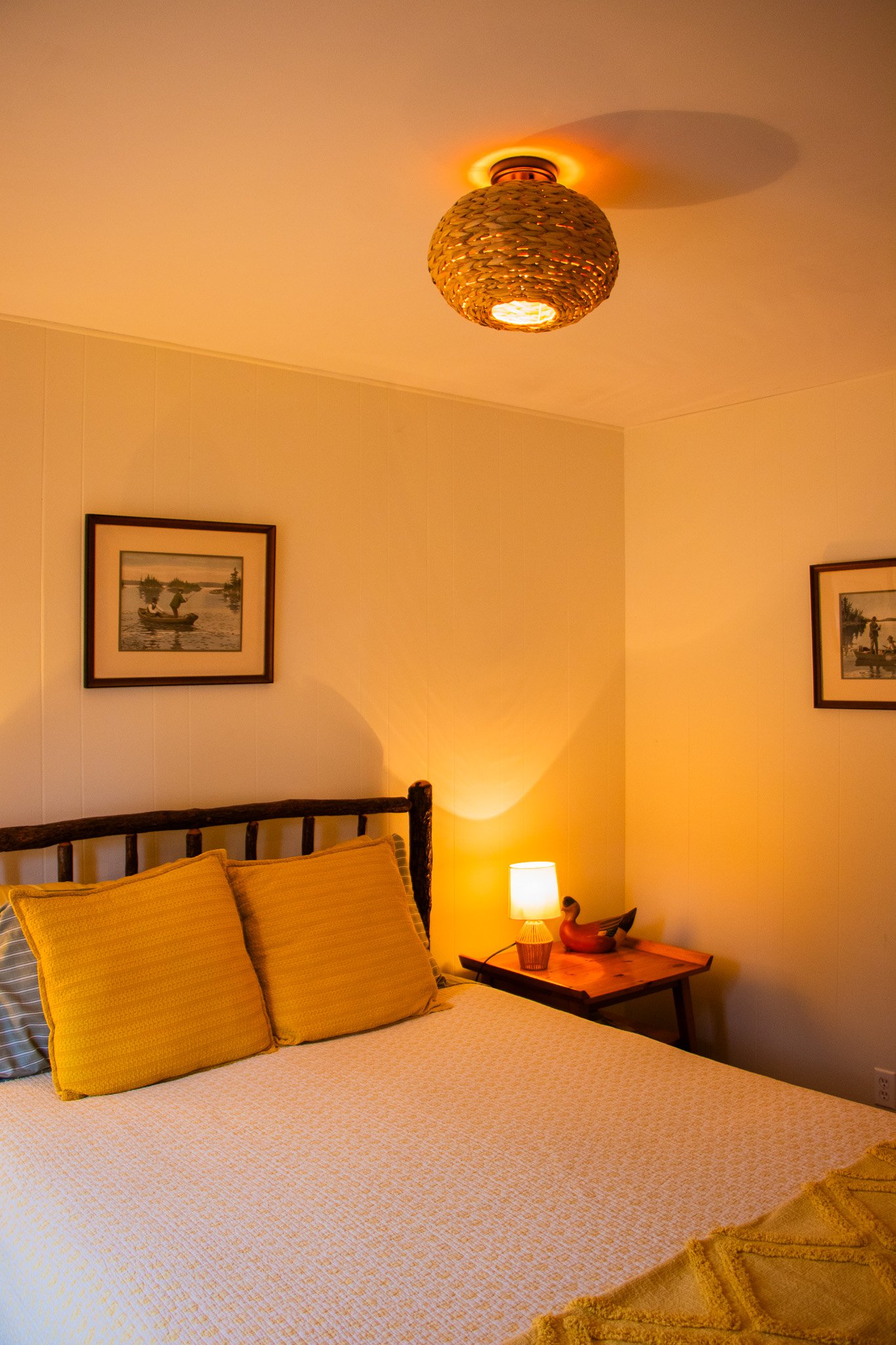 Cozy bedroom with a bed, yellow pillows, a wooden headboard, a nightstand with a lamp and decorative duck figurine, framed artwork on the walls, and warm ambient lighting.