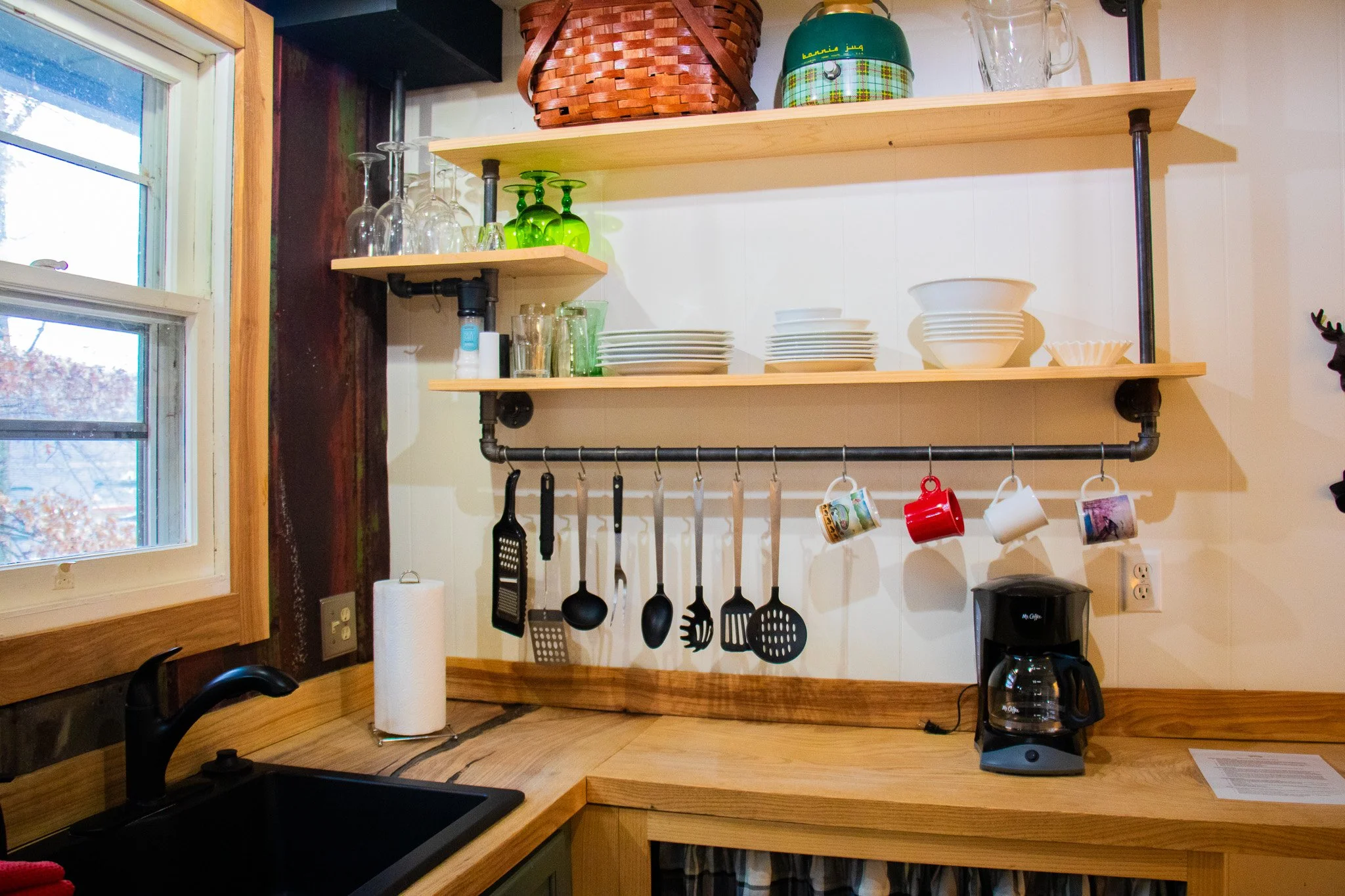 Open kitchen shelf with glassware and dishes, hanging utensils and cups, coffee maker below, window to the left, paper towel on the counter.