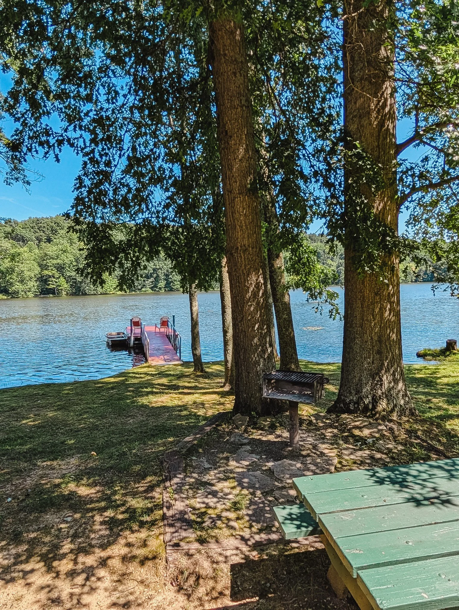 A serene lakeside scene with tall trees providing shade, a wooden dock extending into the water with two chairs on it, a picnic table in the foreground, and lush green hills in the background under a clear blue sky.
