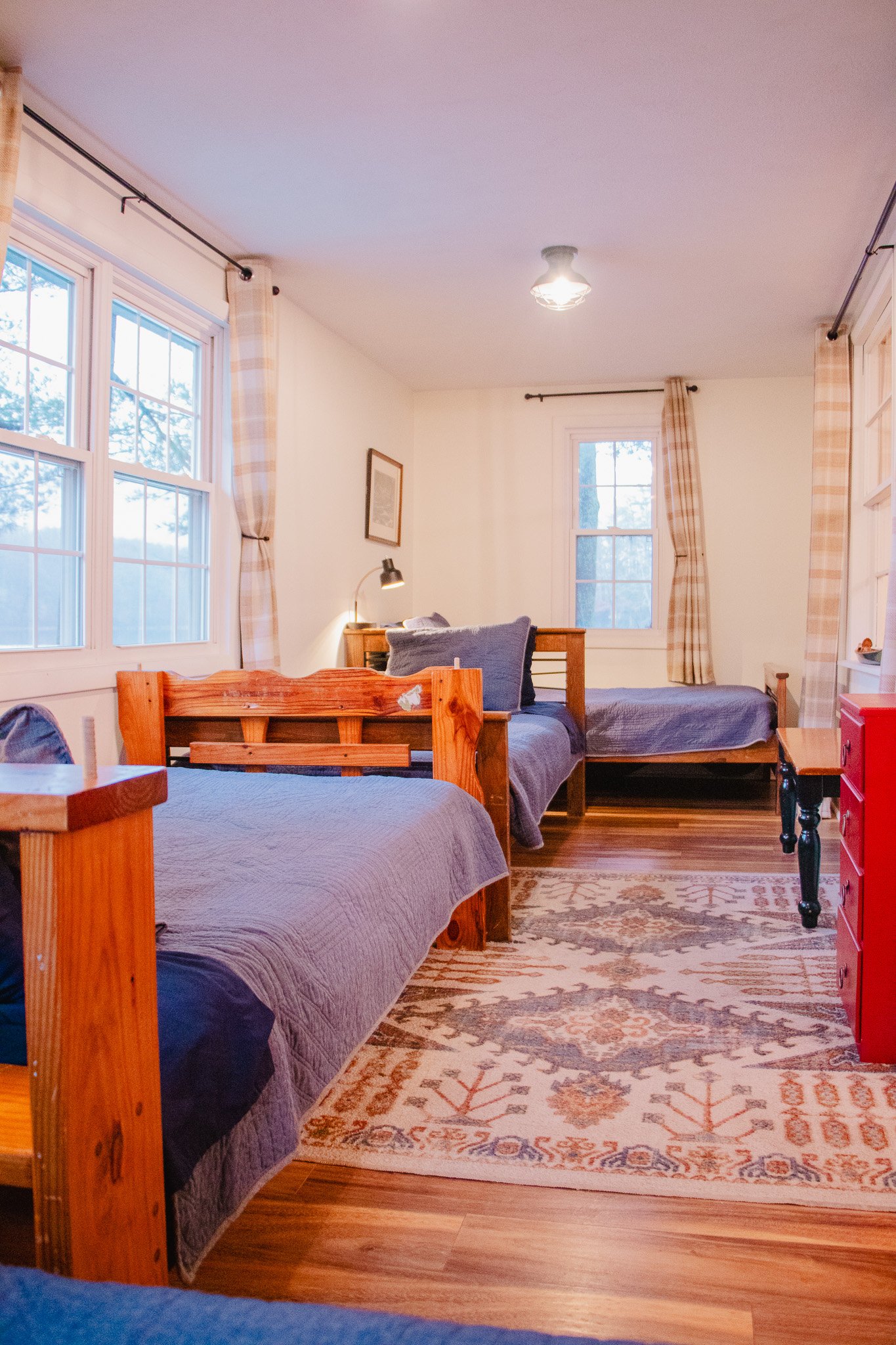 A cozy bedroom with three wooden beds with gray bedding, windows with plaid curtains, a patterned rug, and a red dresser.