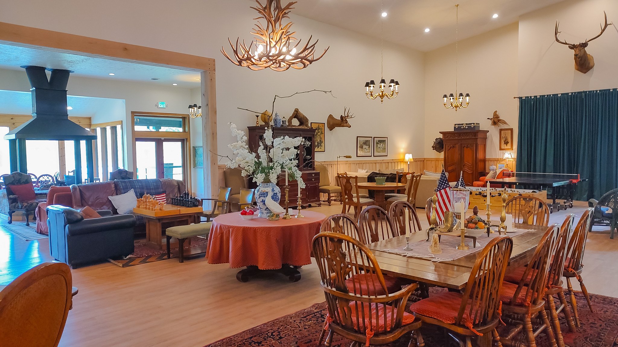 Living and dining area decorated with hunting trophies, chandeliers, and American flags, featuring a tables, sofas, chessboard, ping pong table, and floral centerpiece.