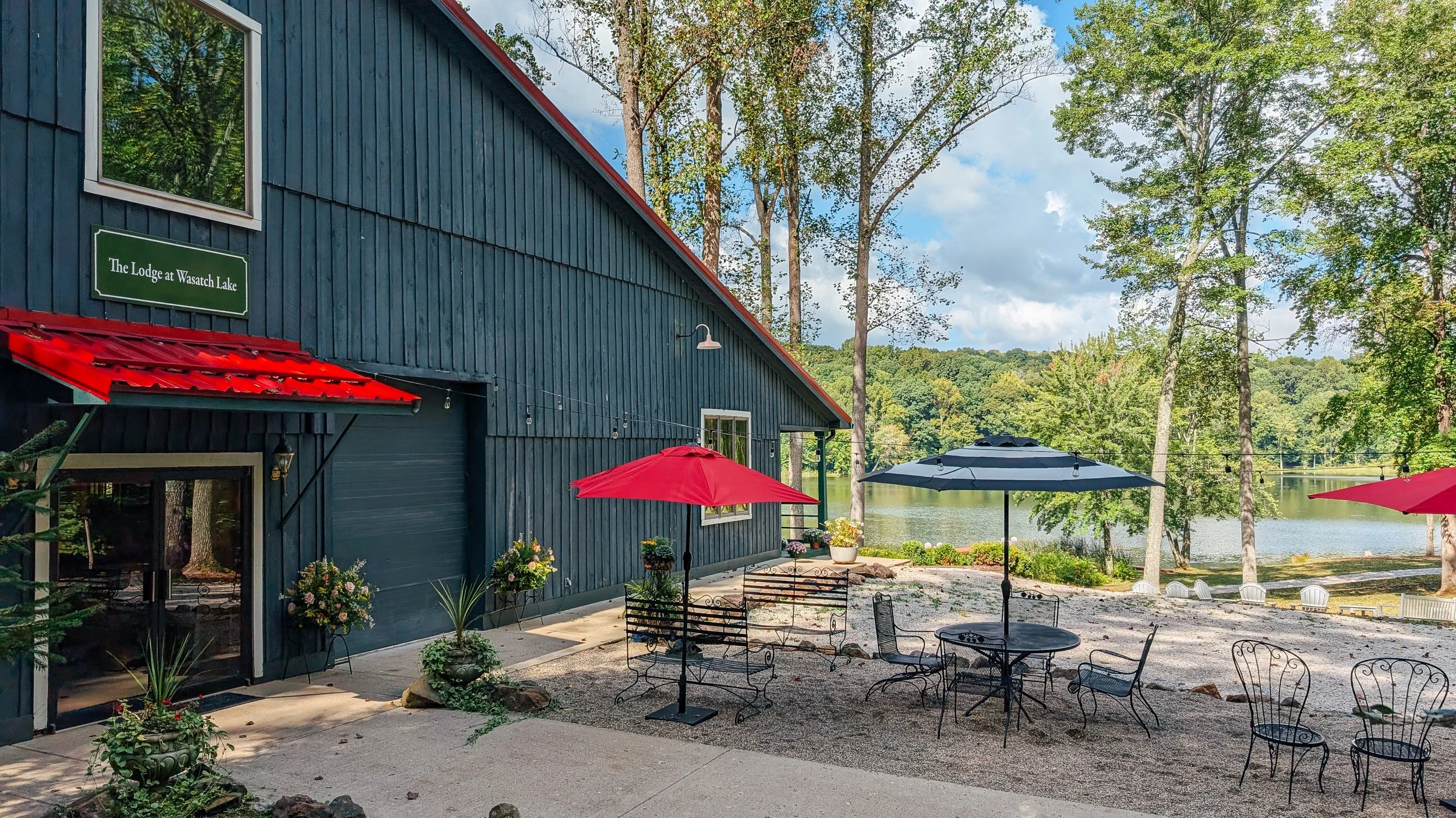 Outdoor seating area with black wrought iron tables and chairs, red and dark gray umbrellas, near a blue lakeside building with a sign that reads 'The Lodge at Wasatch Lake.' In the background, there are tall trees, a lake, and a partly cloudy sky.