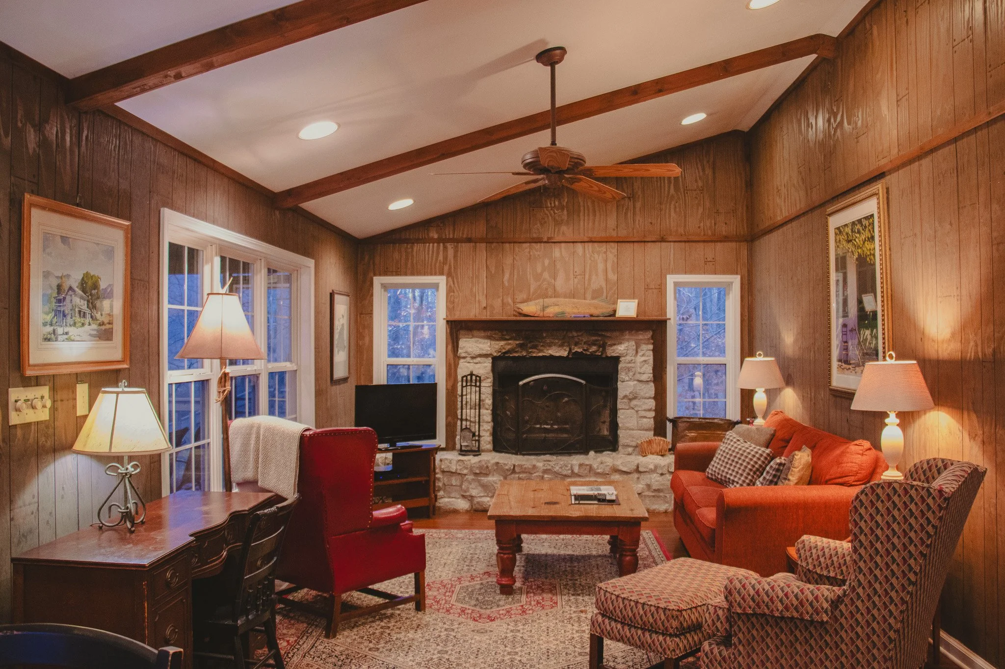 Cozy living room with wood-paneled walls, a stone fireplace, a TV, a red armchair, a plaid armchair with an ottoman, a sofa with pillows, lamps, and framed artwork.