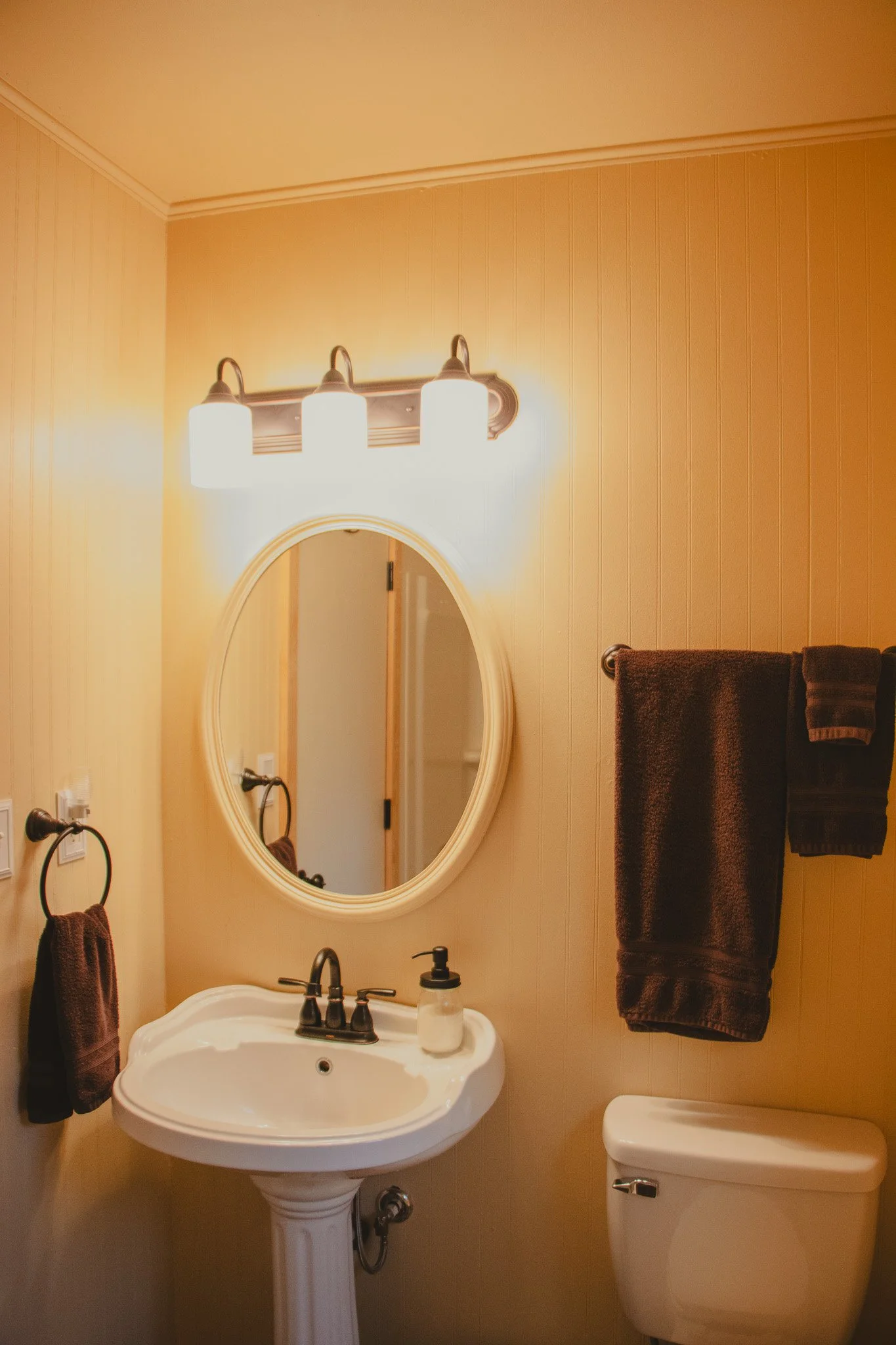 A small bathroom with a pedestal sink, oval mirror, wall-mounted light fixture, dark towels on a towel rack, and a toilet.