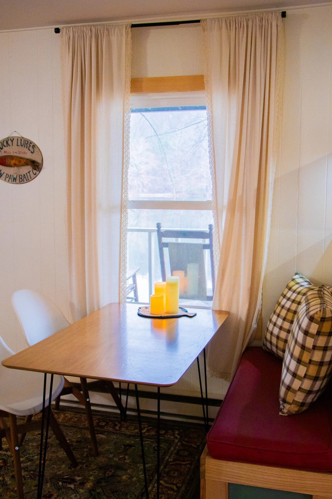 A cozy dining nook with a wooden table, white chairs, a bench with plaid pillows, and a window with beige curtains, decorated with candles on the table, overlooking a snowy outdoor scene.