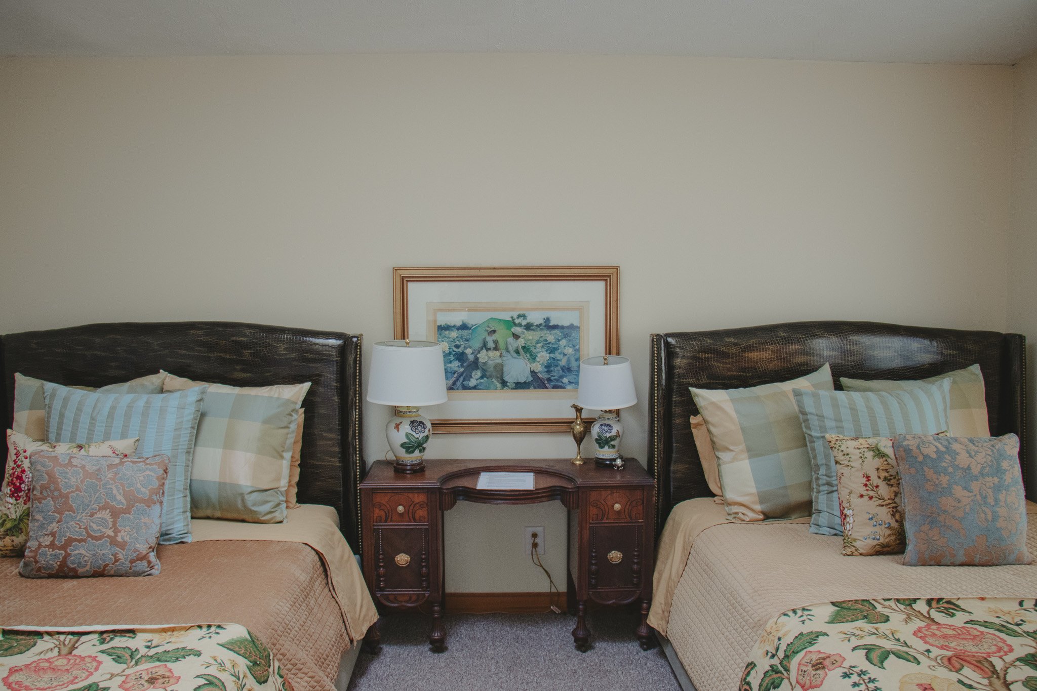 Two beds with dark wooden headboards and beige bedding, pillows with floral and striped patterns, a wooden nightstand with lamps and a framed picture between the beds.