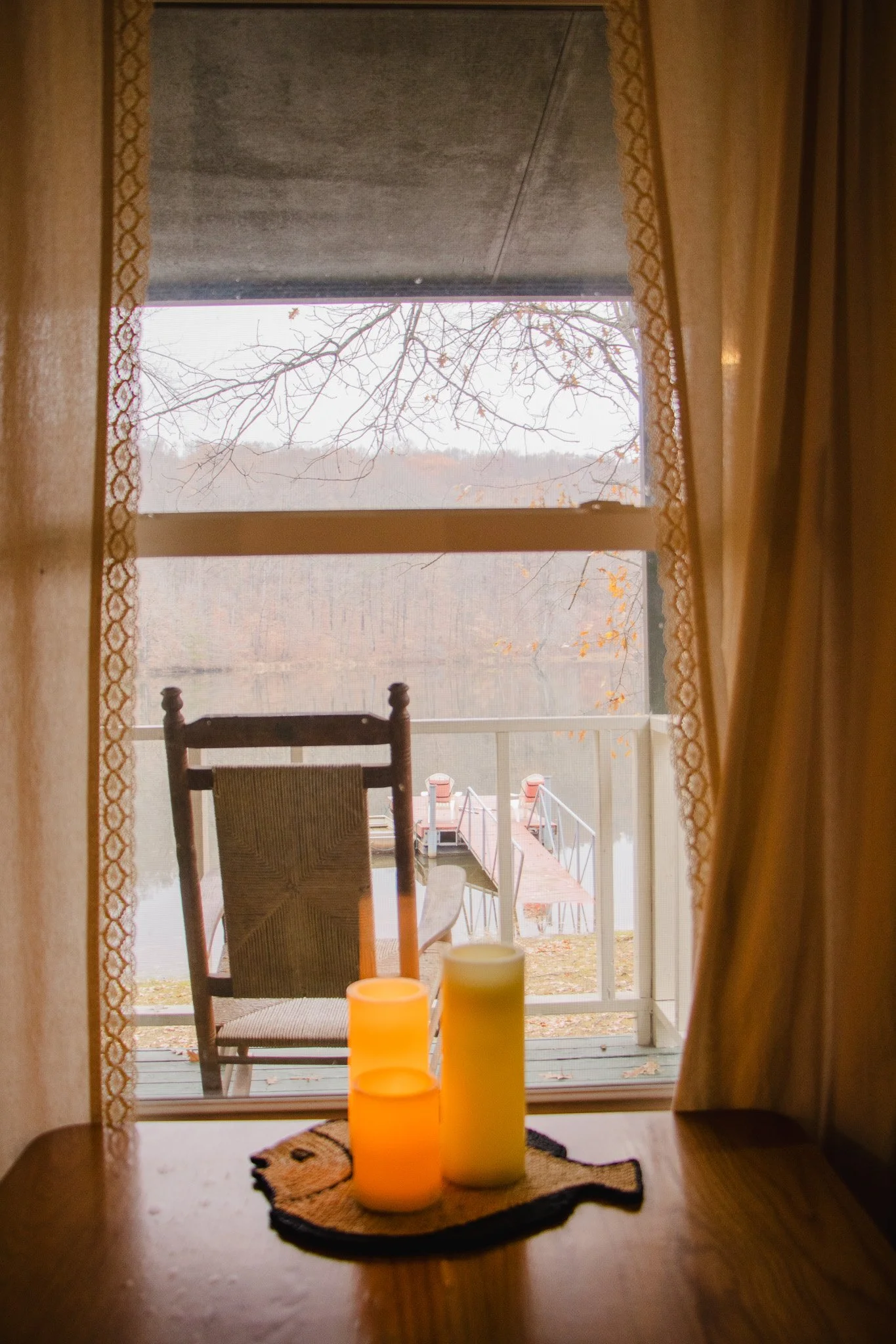 A view through a window showing a lake, trees with fall foliage, a wooden dock, and candles on a table inside.