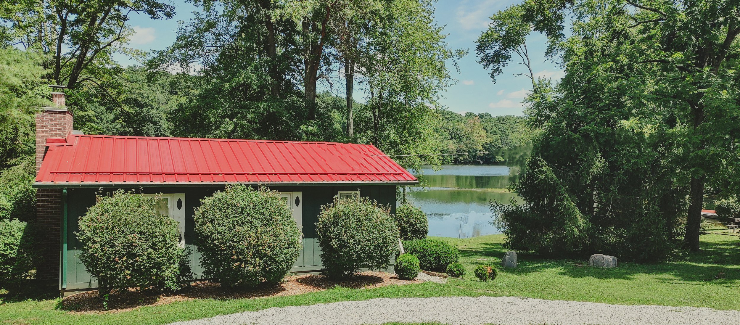 A small green house with a red metal roof, surrounded by shrubs and trees, with a lake in the background under a blue sky.