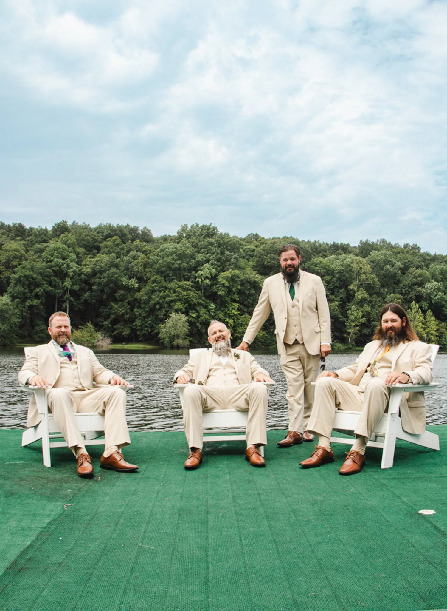 Four men in beige suits sitting and standing on a green platform by a lake with trees in the background, during daytime.