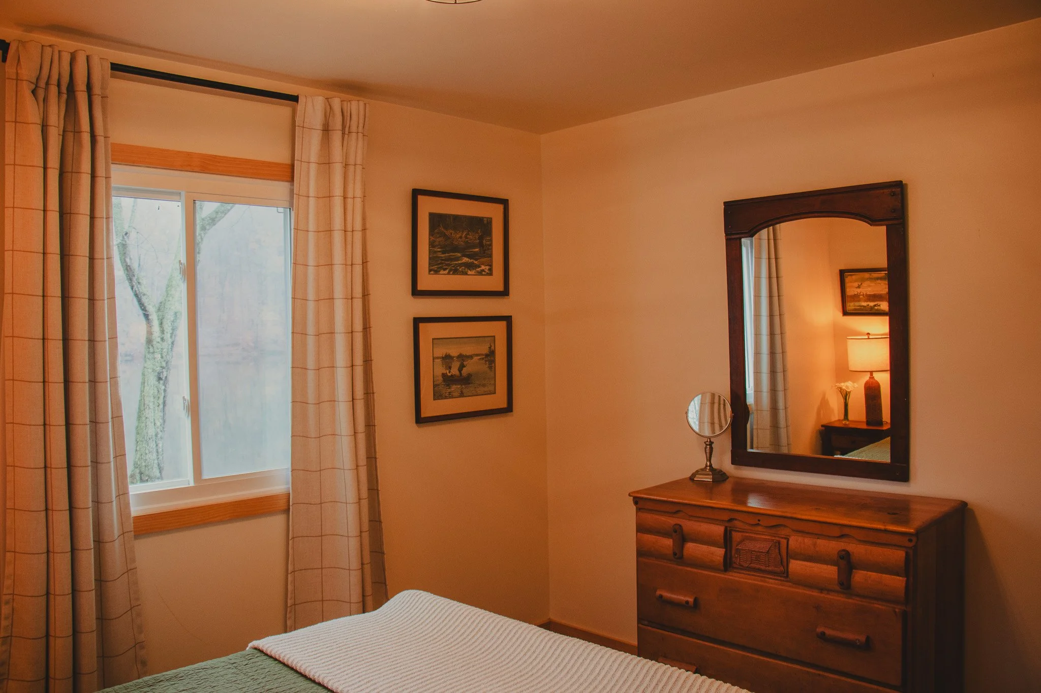 A cozy bedroom with a window, beige curtains, framed landscape pictures on the wall, a wooden dresser with a mirror, and a lamp on a small nightstand in the reflected room.