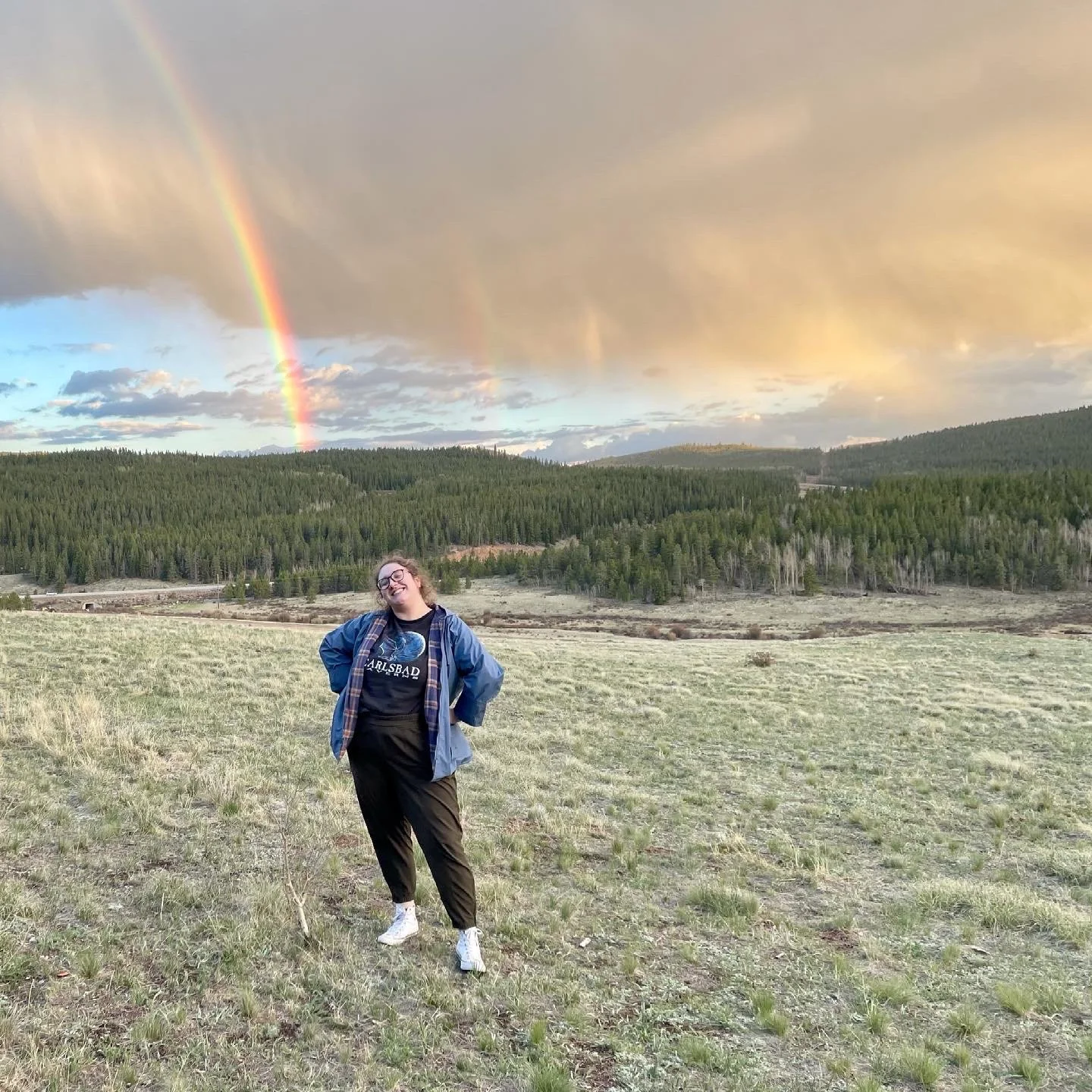 A person standing in a grassy field with a forested hill and rainbow in the sky behind them. The person is smiling, wearing glasses, a blue jacket, and a black t-shirt with white text.