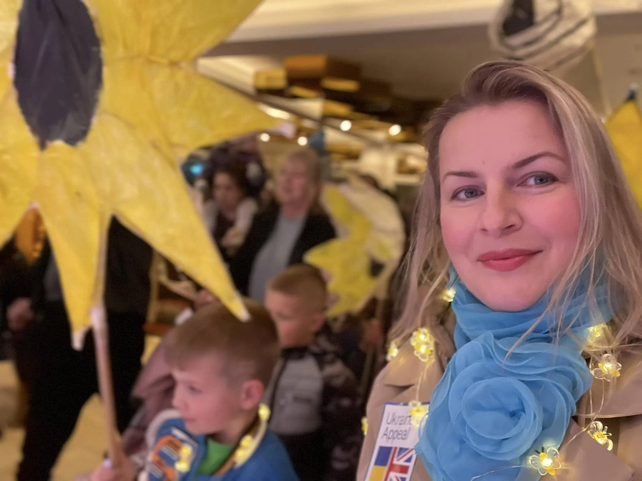 Woman wearing a Ukraine Appeal badge and flower lights, surrounded by children holding bright yellow star decorations at a community event