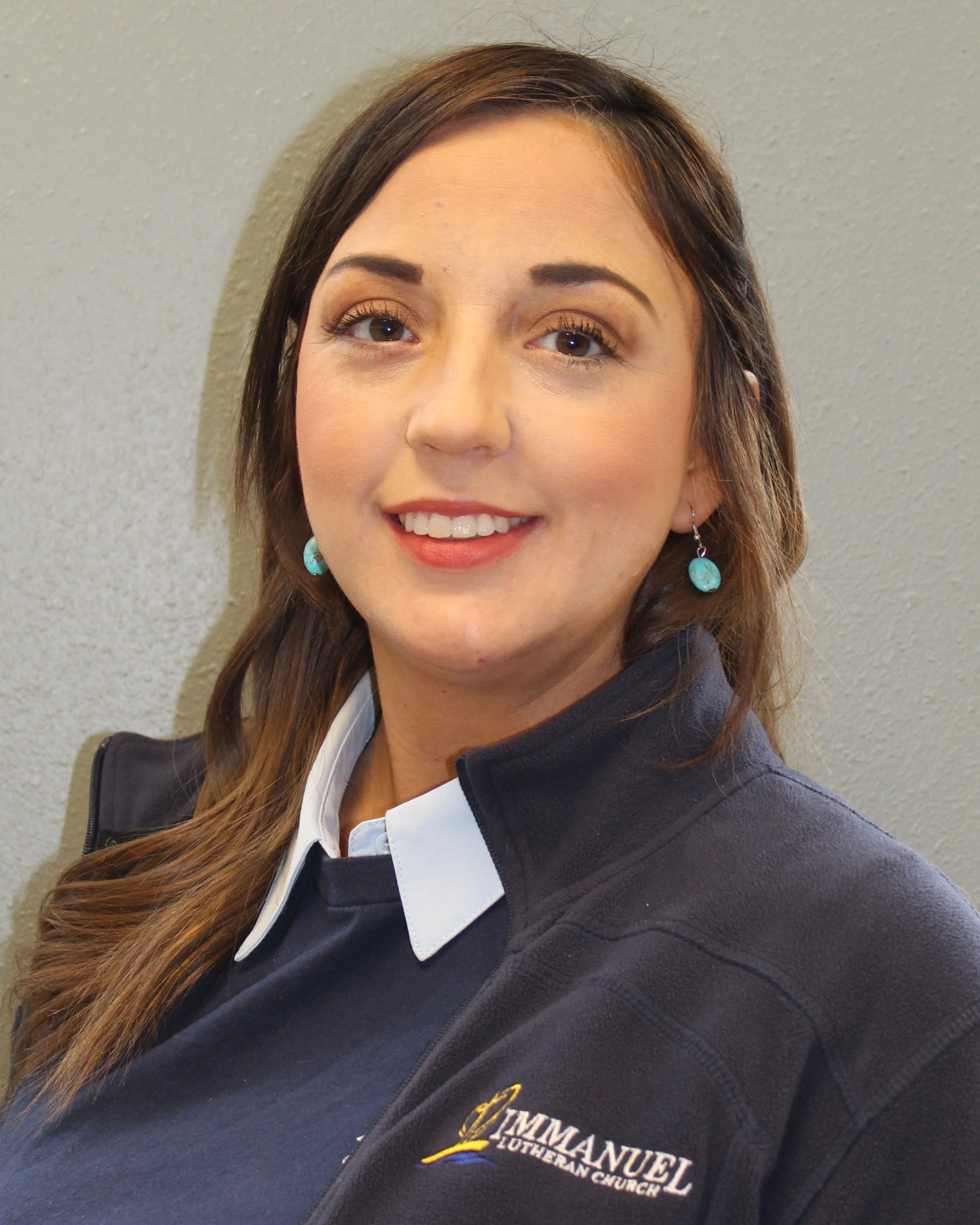 Close-up of a woman smiling, with brown hair and turquoise earrings, wearing a black fleece jacket with embroidered logo of Immanuel Lutheran Church.