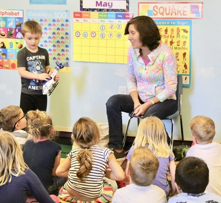 A woman sits on a chair in front of a group of young children inside a classroom. One boy is standing near her, holding a book. The children are sitting on the floor, facing the woman. The classroom has educational posters, including a calendar for May and alphabet and number charts.
