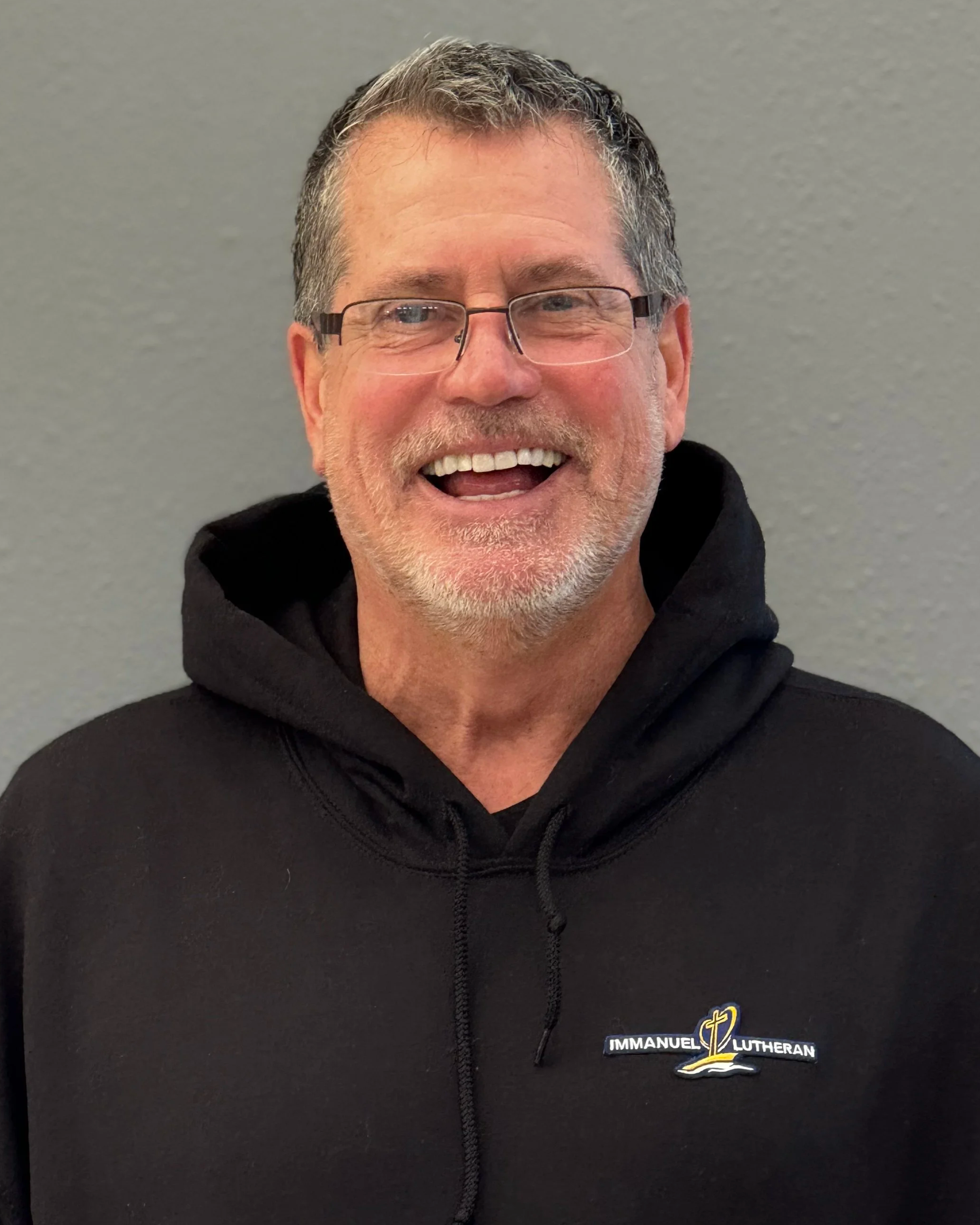 Smiling middle-aged man with glasses and gray hair wearing a black hoodie with an 'Immanuel Lutheran' logo, standing against a plain gray background.