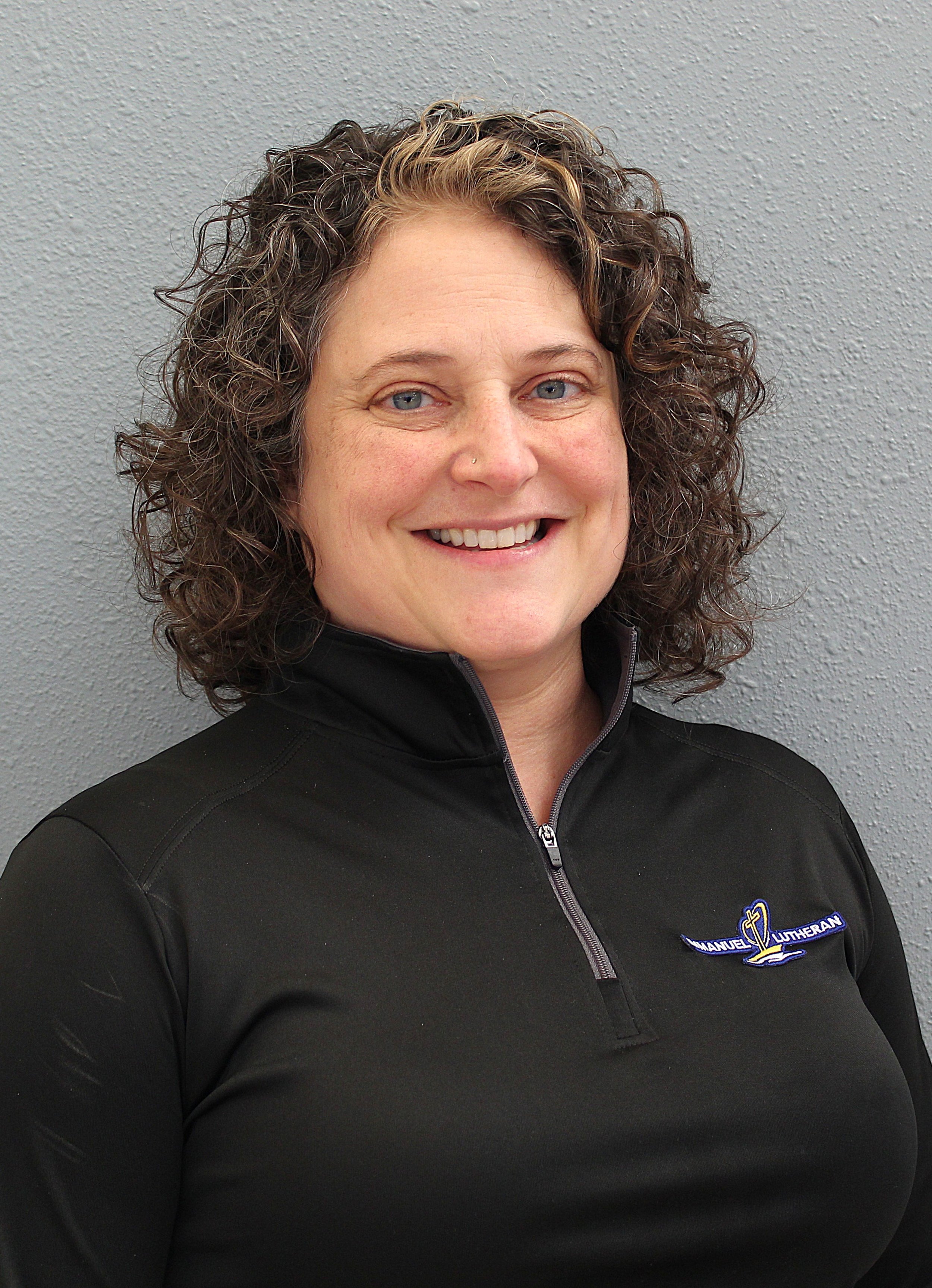A smiling woman with curly hair wearing a black jacket with a church logo on it, standing against a grey textured wall.