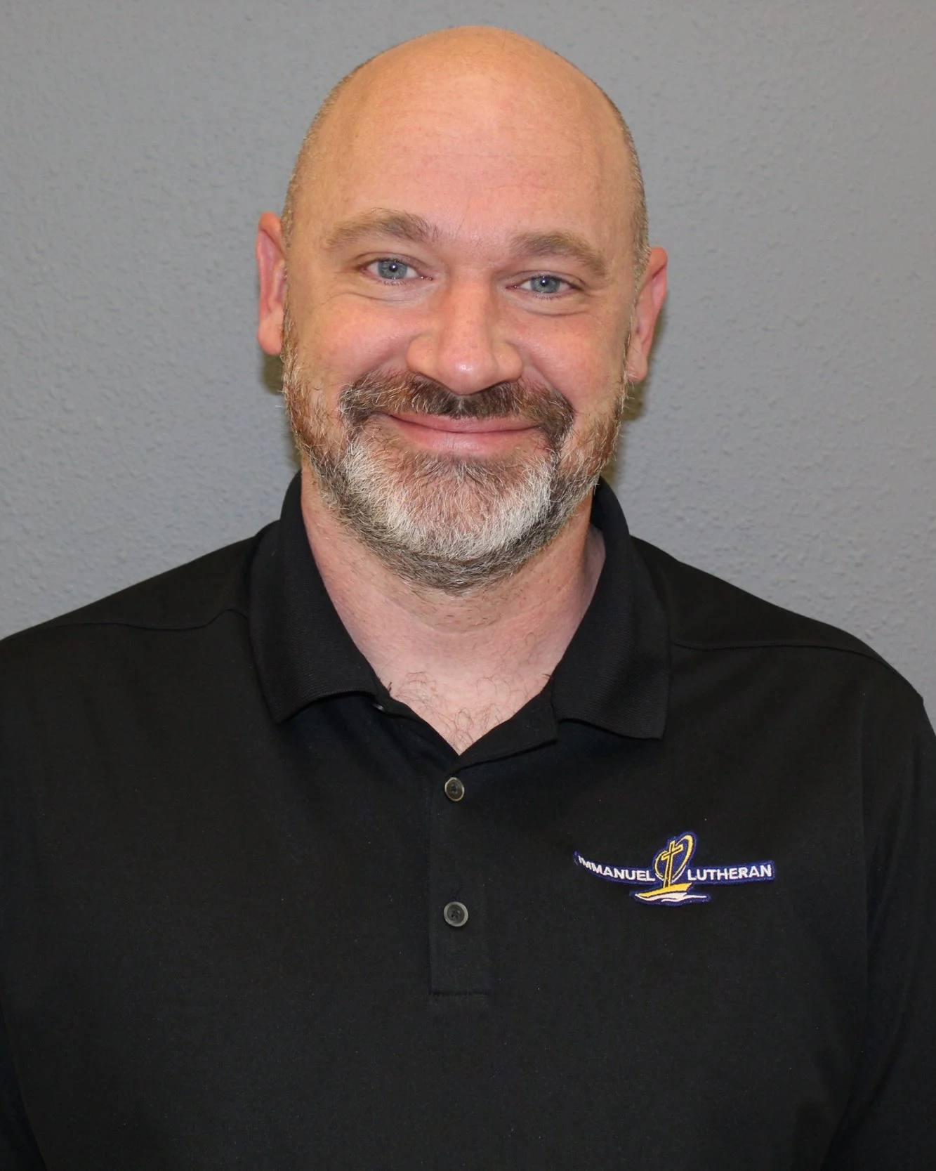 A smiling man with a beard and bald head wearing a black shirt with an Emmanuel Lutheran logo.