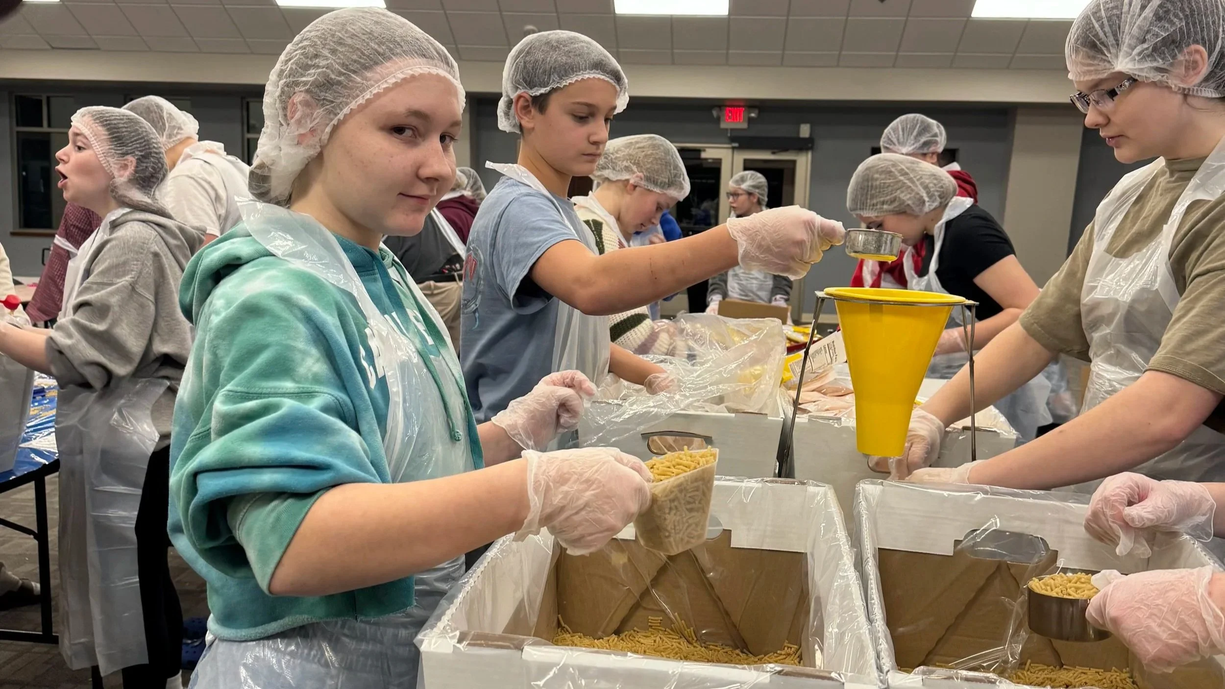 Group of children making pasta in a large industrial kitchen, wearing hairnets, gloves, and aprons.