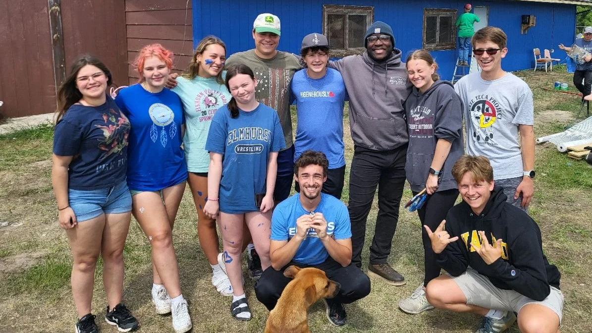 Group of teenagers and young adults outdoors, some with face paint, smiling, standing close together, with two men in the middle, one crouching with a dog, and a young man making a hand gesture. Background includes a building and other people.
