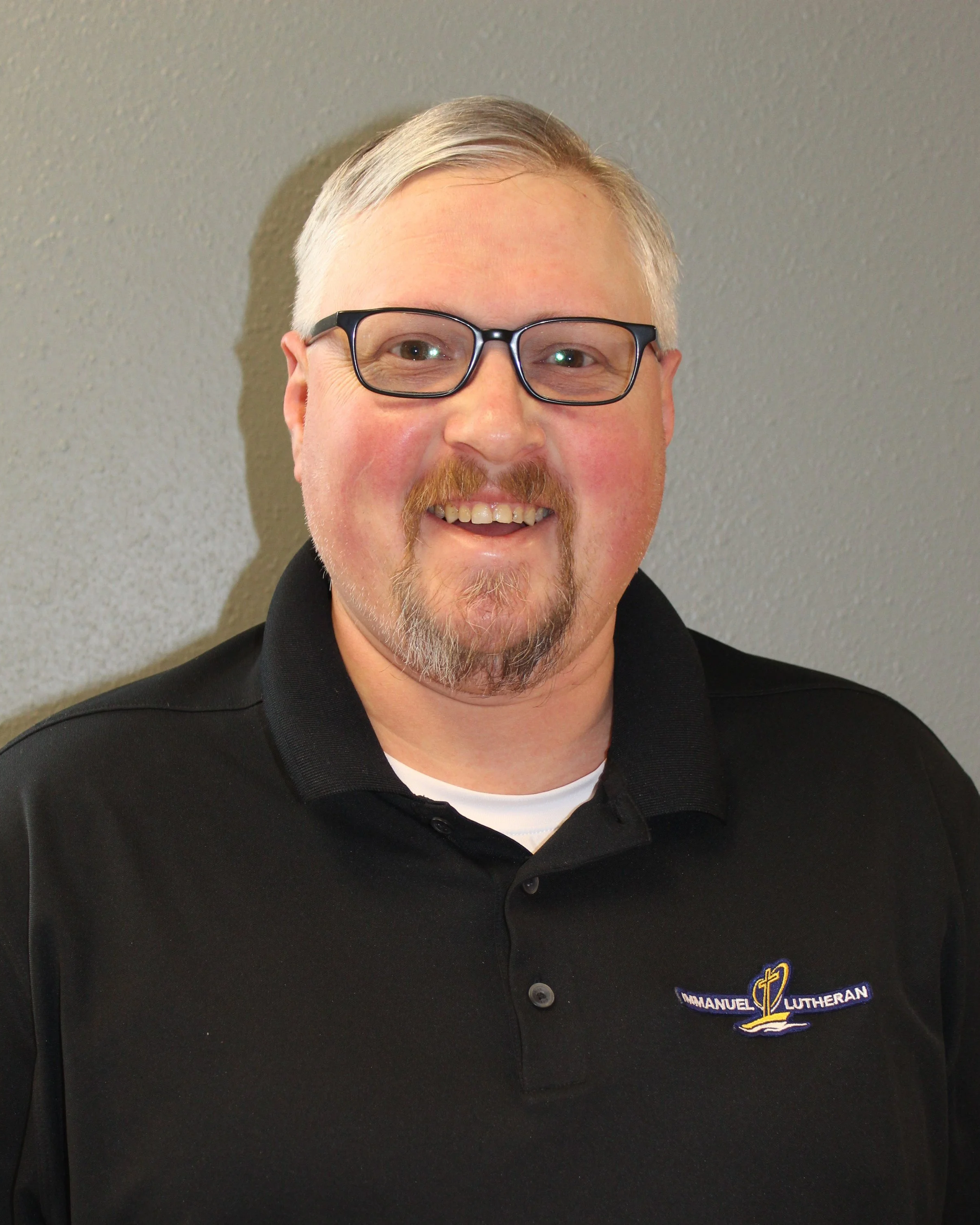 A smiling man with glasses, light-colored hair, and a goatee wearing a black polo shirt with a logo that says "Emmanuel Lutheran" and a cross design on it.