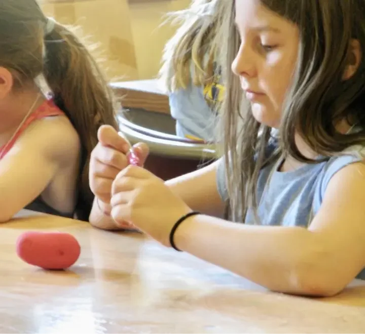 A young girl with long brown hair, wearing a blue shirt, sitting at a table, peeling a pink plastic egg while two other children are partially visible nearby, in a classroom or similar setting.