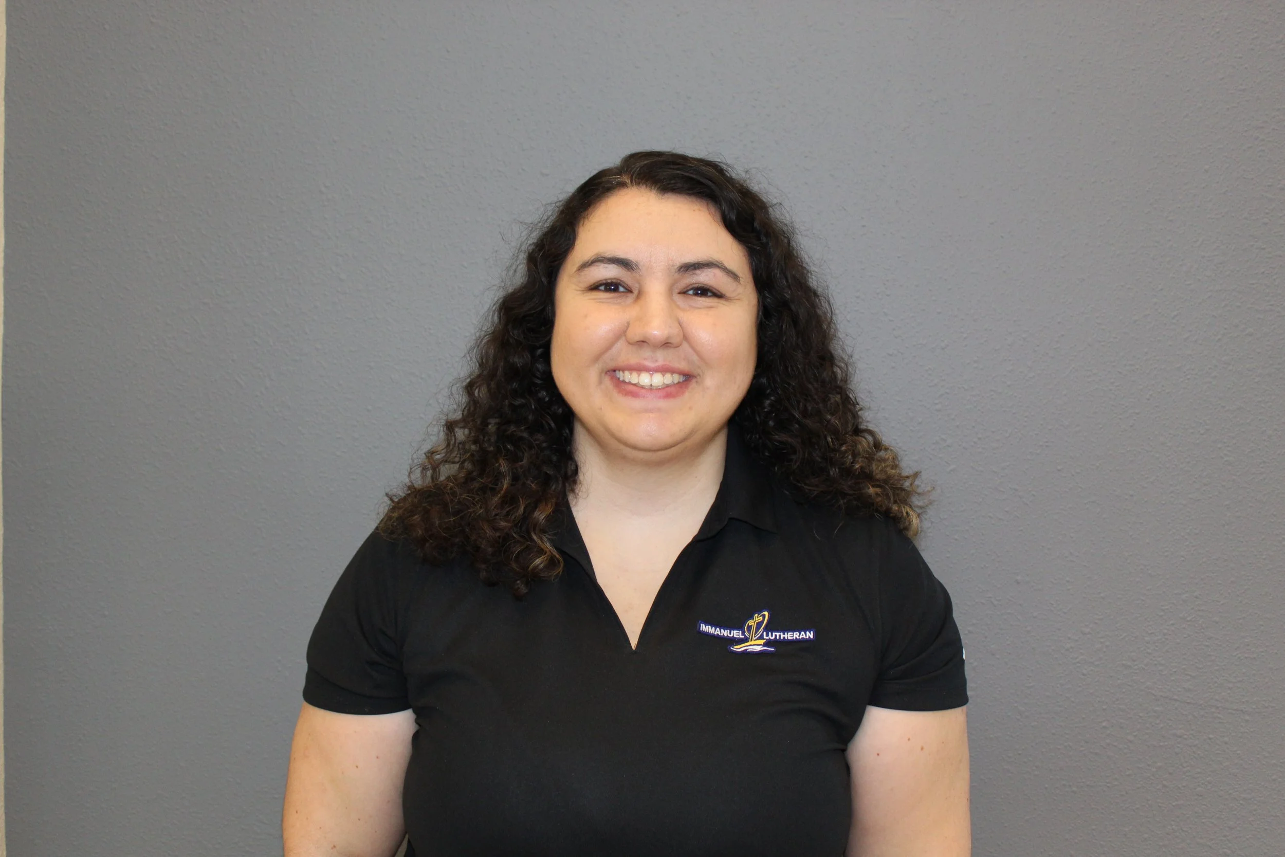 A young woman with curly brown hair is smiling, wearing a black polo shirt with 'Immanuel Lutheran' embroidered on it, standing against a gray textured wall.