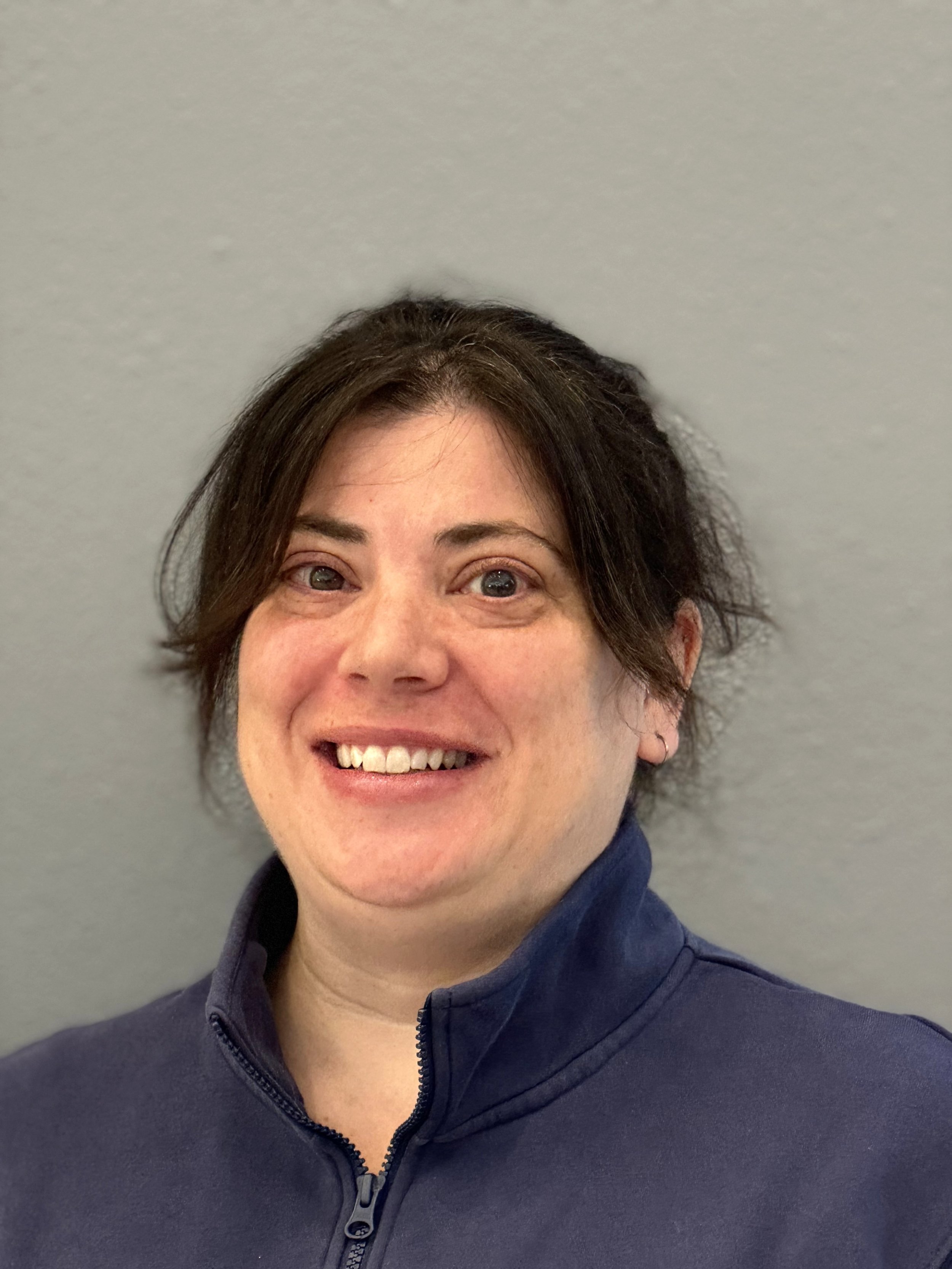 Close-up of a woman with short brown hair, smiling, wearing a navy blue zip-up jacket against a plain light gray background.