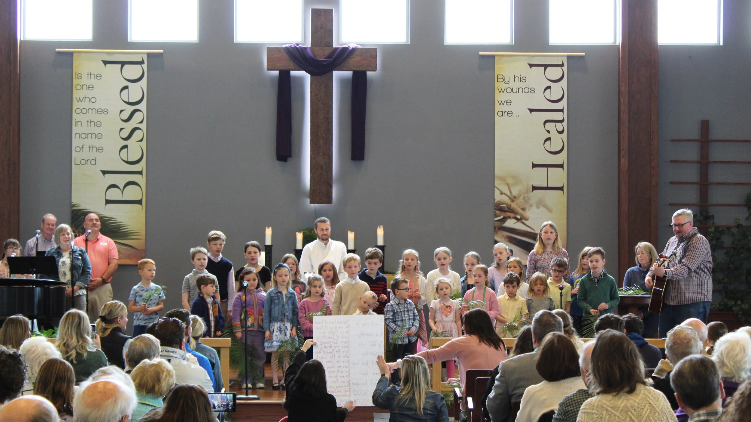 A congregation of children and adults gathered in a church with a large wooden cross and banners that read "Blessed" and "His wounds we are..." The children are standing on stage singing or performing, while the audience watches. Some adults are playing instruments, including a man with a guitar.