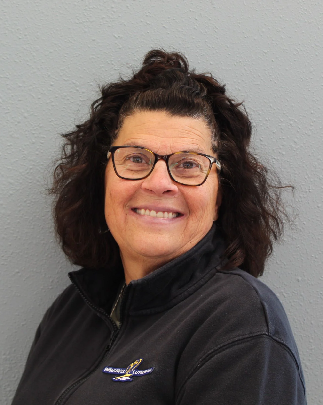 Smiling woman with curly brown hair tied back, wearing glasses and a black zip-up jacket, with a gray wall background.