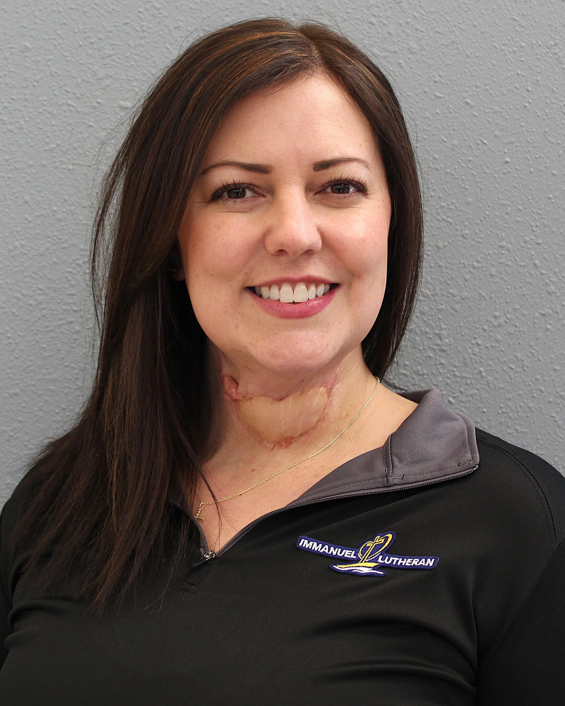 A woman with long brown hair smiling at the camera, wearing a black jacket with an 'Immanuel Lutheran' logo and a gold necklace, standing against a gray textured wall.