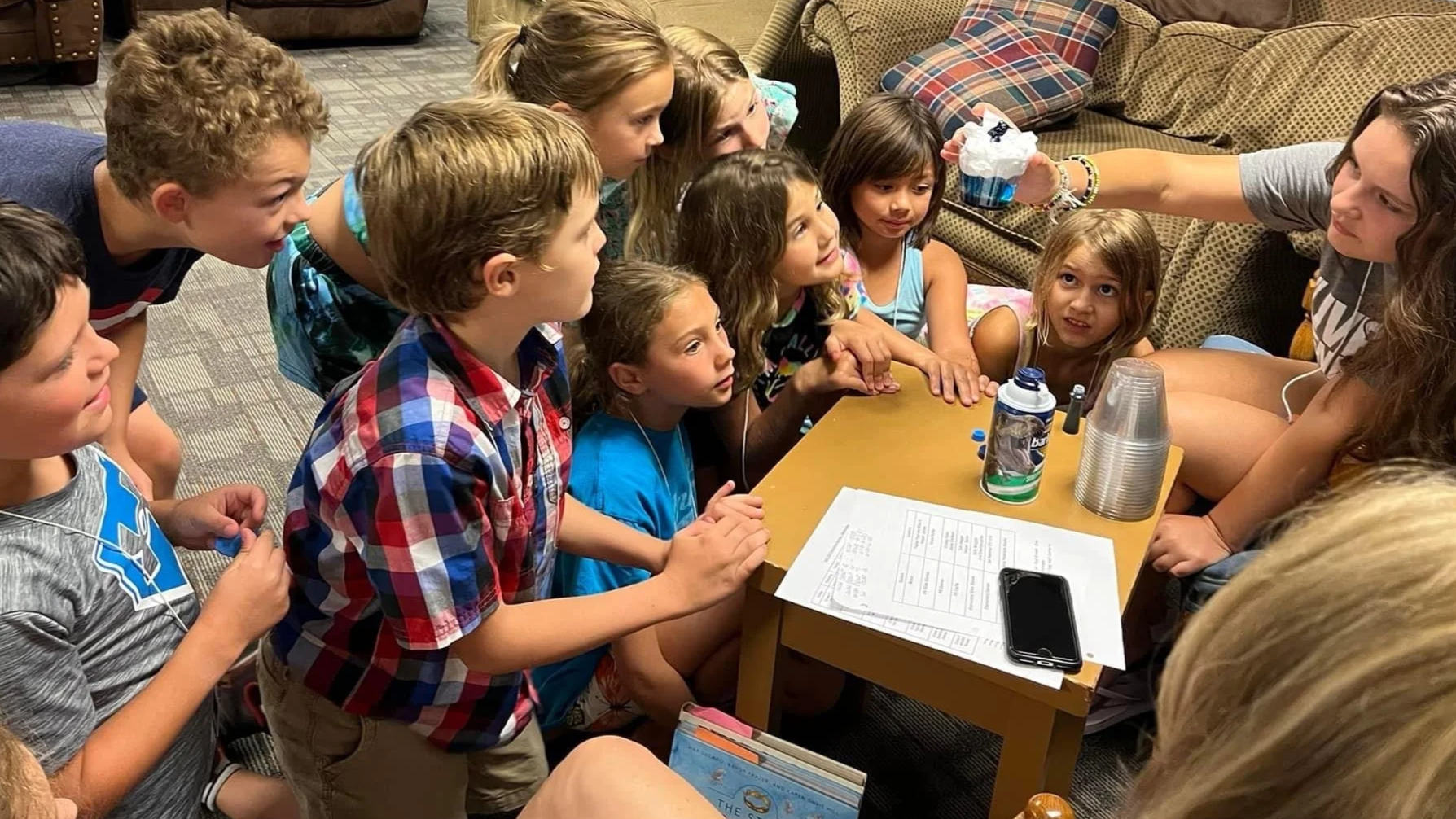Group of children gathered around a table, with an adult woman distributing items, possibly at a party or event.