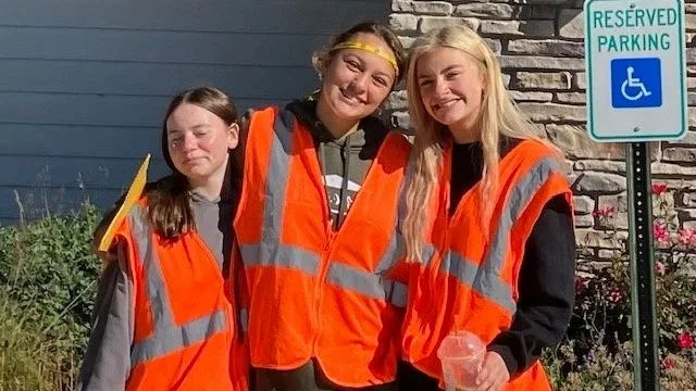 Three young girls wearing orange safety vests standing outside near a reserved parking sign, with one holding a clear cup.